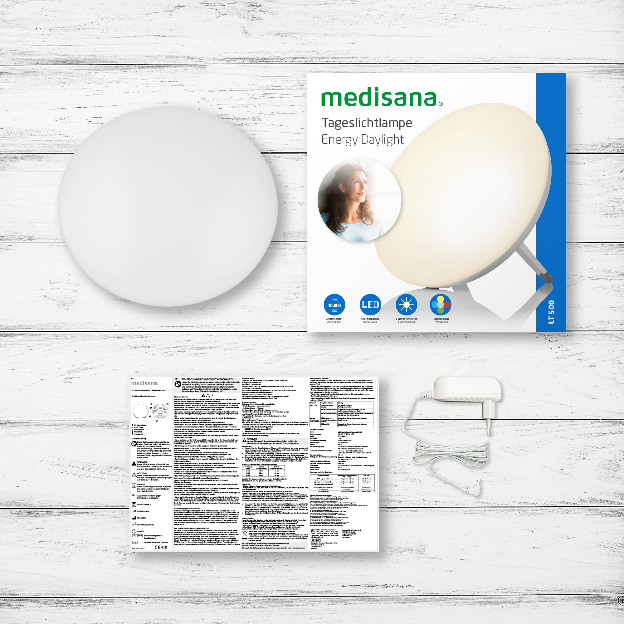 Eine weiße Lichttherapielampe und -box, mit Handbuch und Netzteil, auf einem Holztisch.