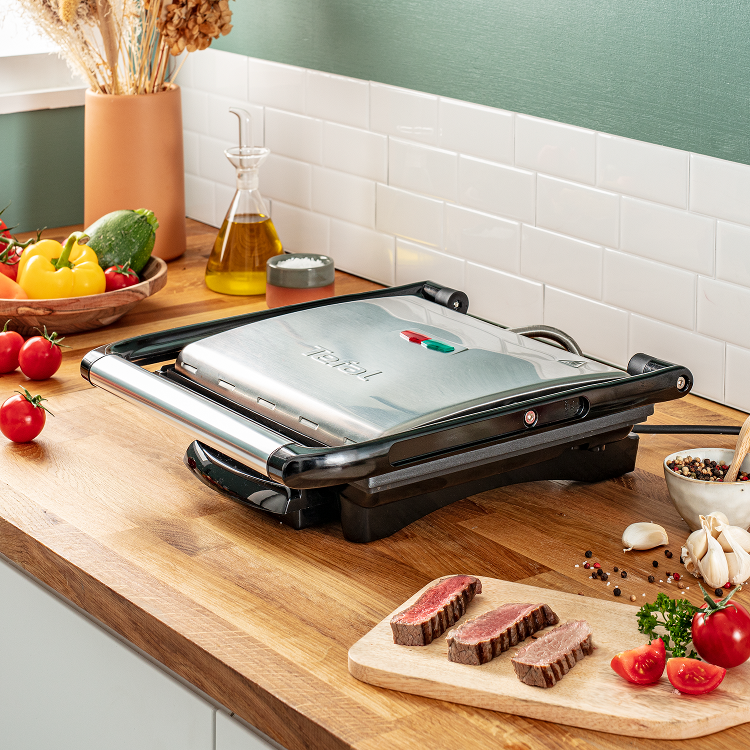 Parrilla Tefal sobre superficie de madera con ingredientes. Bistec, tomates, ajo y hierbas visibles.