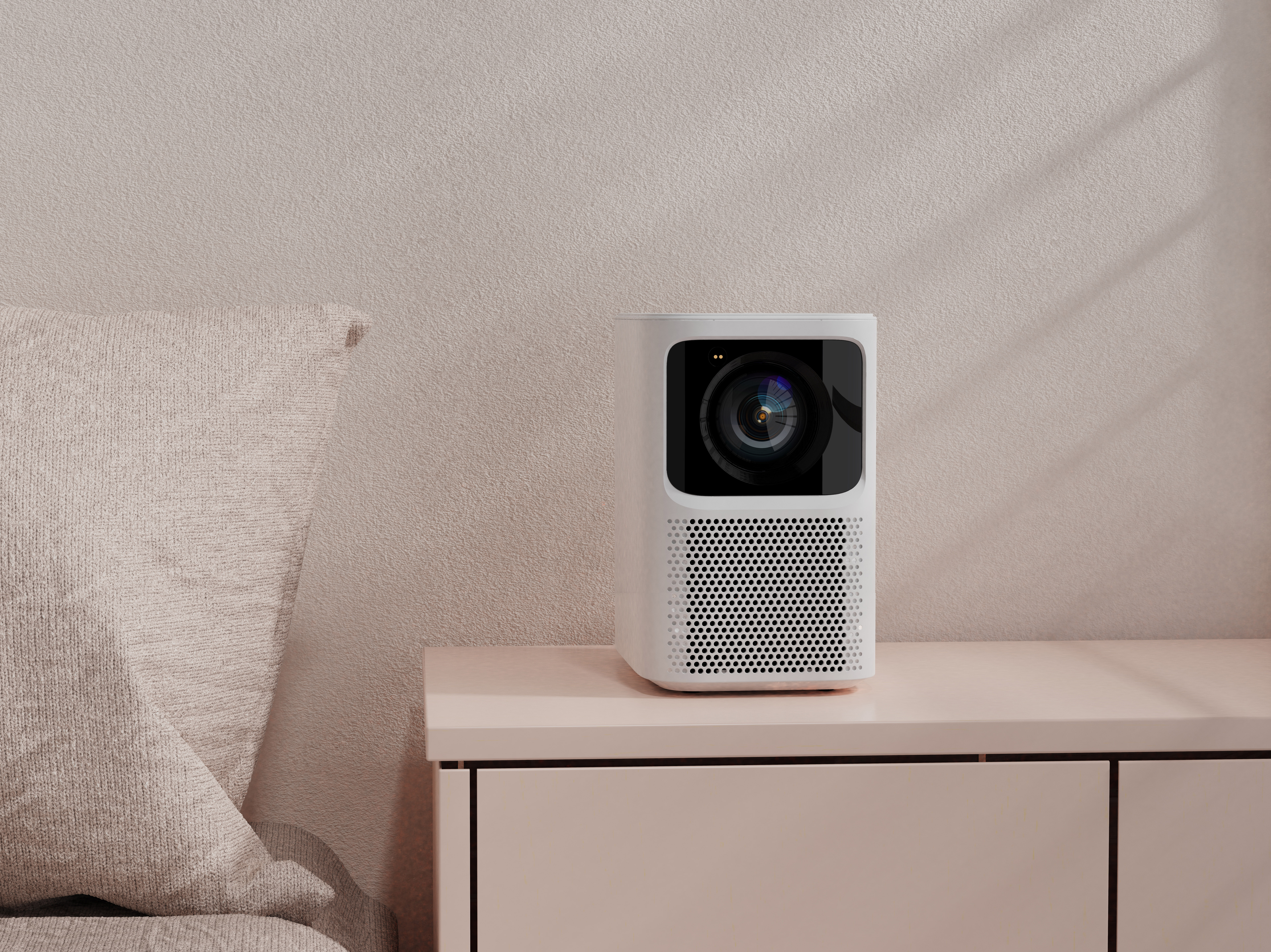 A white projector on a pink table, with a beige pillow and wall in the background.