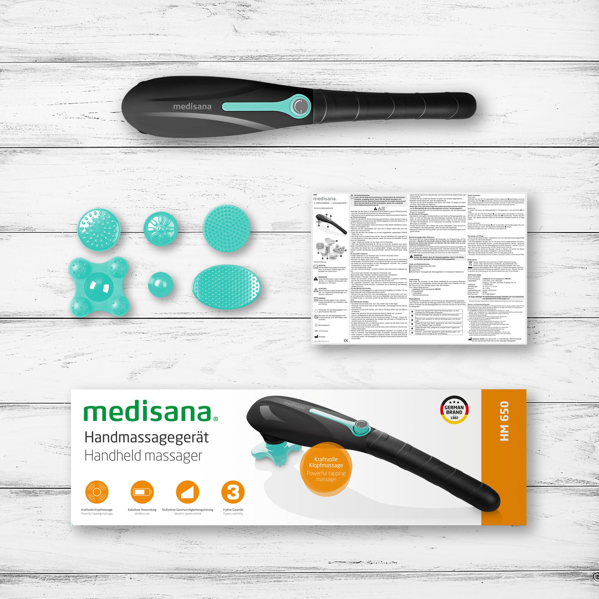 Ein Handmassagegerät mit Zubehör und Verpackung. Das Massagegerät ist schwarz und blaugrün. Weißer Holzuntergrund.