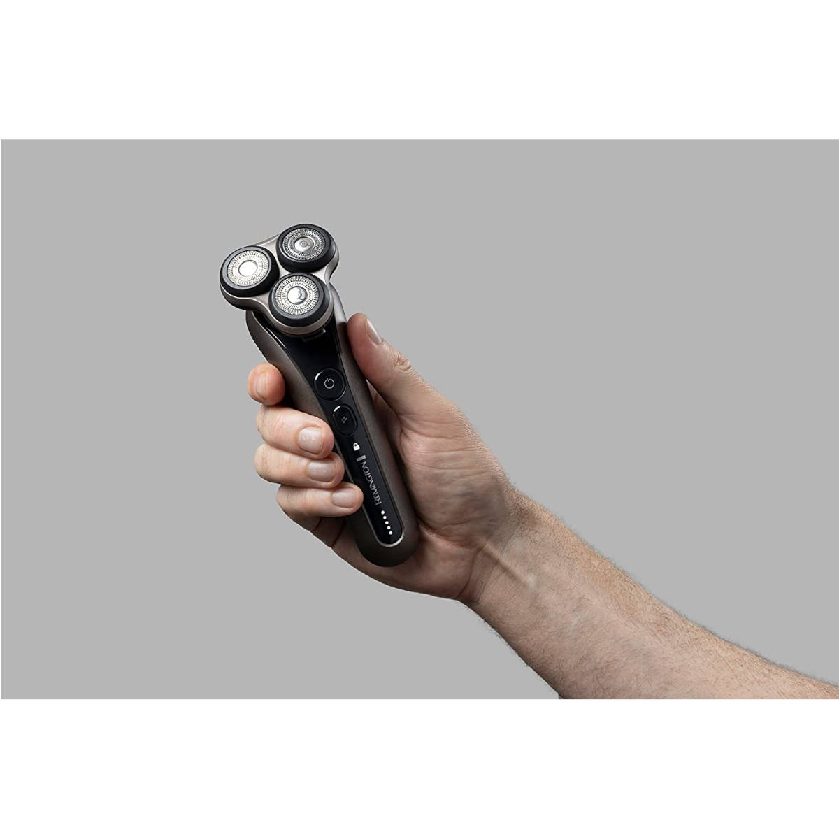 Mann hält einen Remington-Rasierer. Der Rasierer ist dunkelgrau mit drei Rotationsköpfen. Grauer Hintergrund.