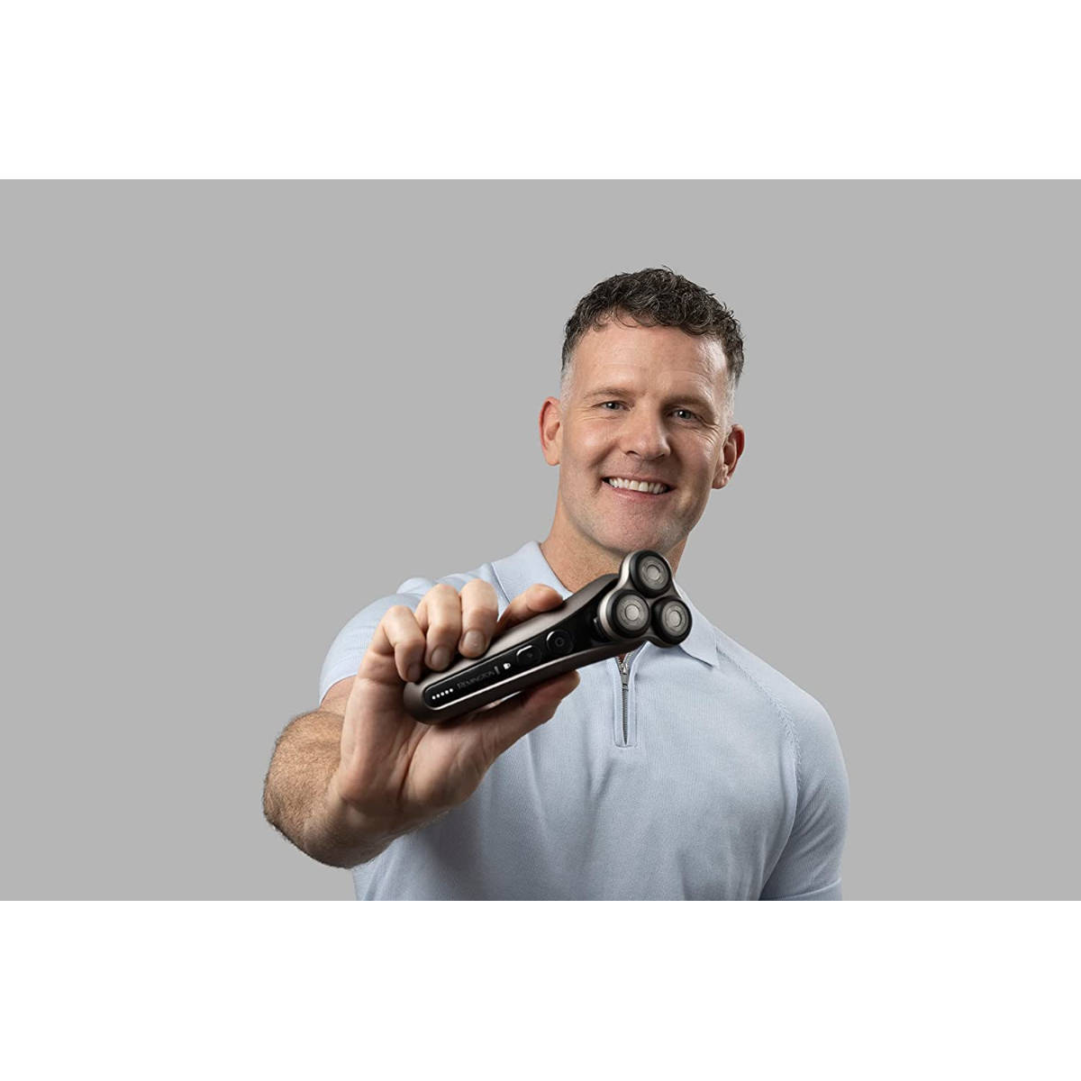 Ein lächelnder Mann, der einen Elektrorasierer in der Hand hält, vor grauem Hintergrund.
