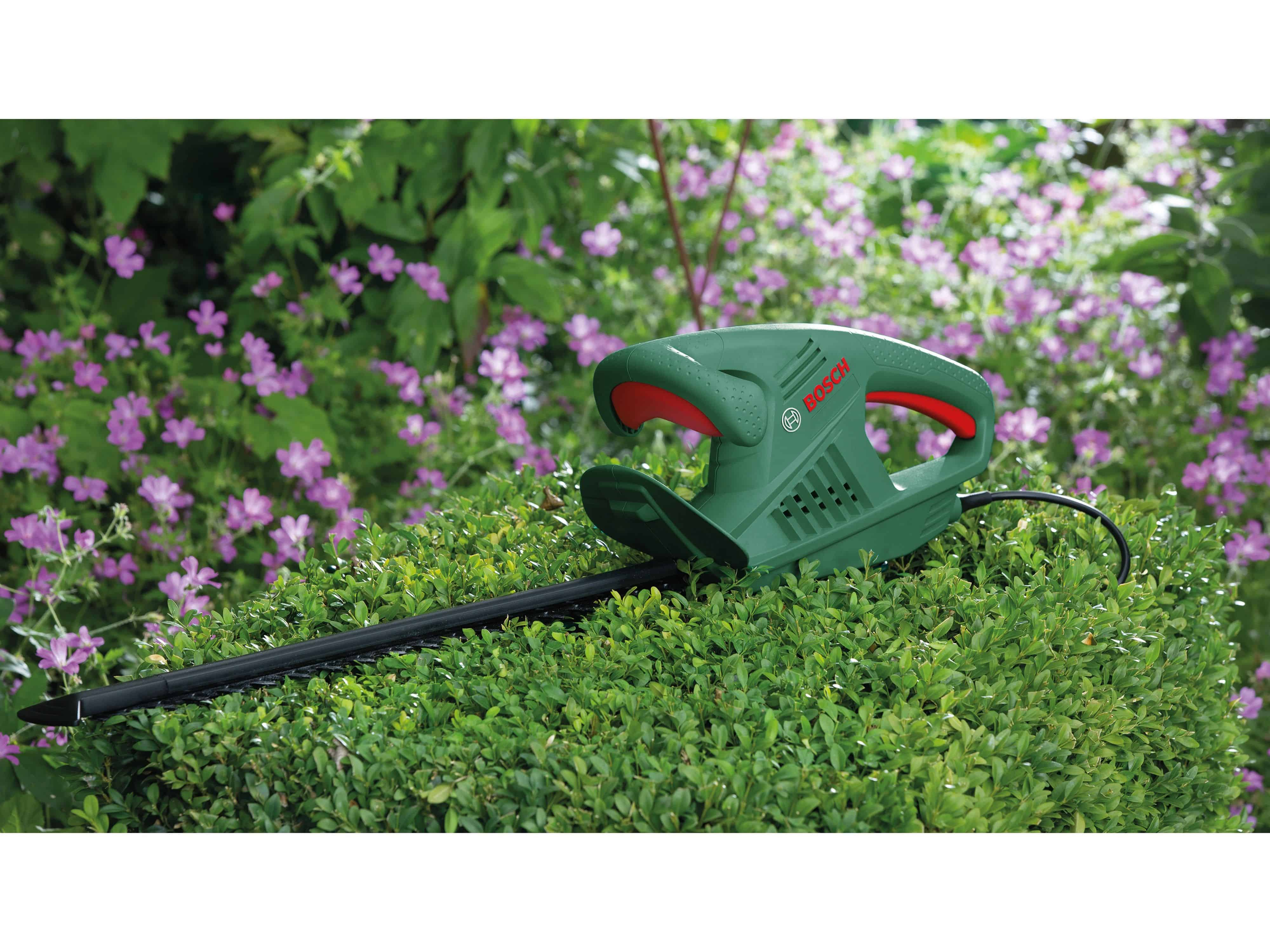 Un taille-haie Bosch vert et noir sur un buisson vert avec des fleurs roses en arrière-plan.