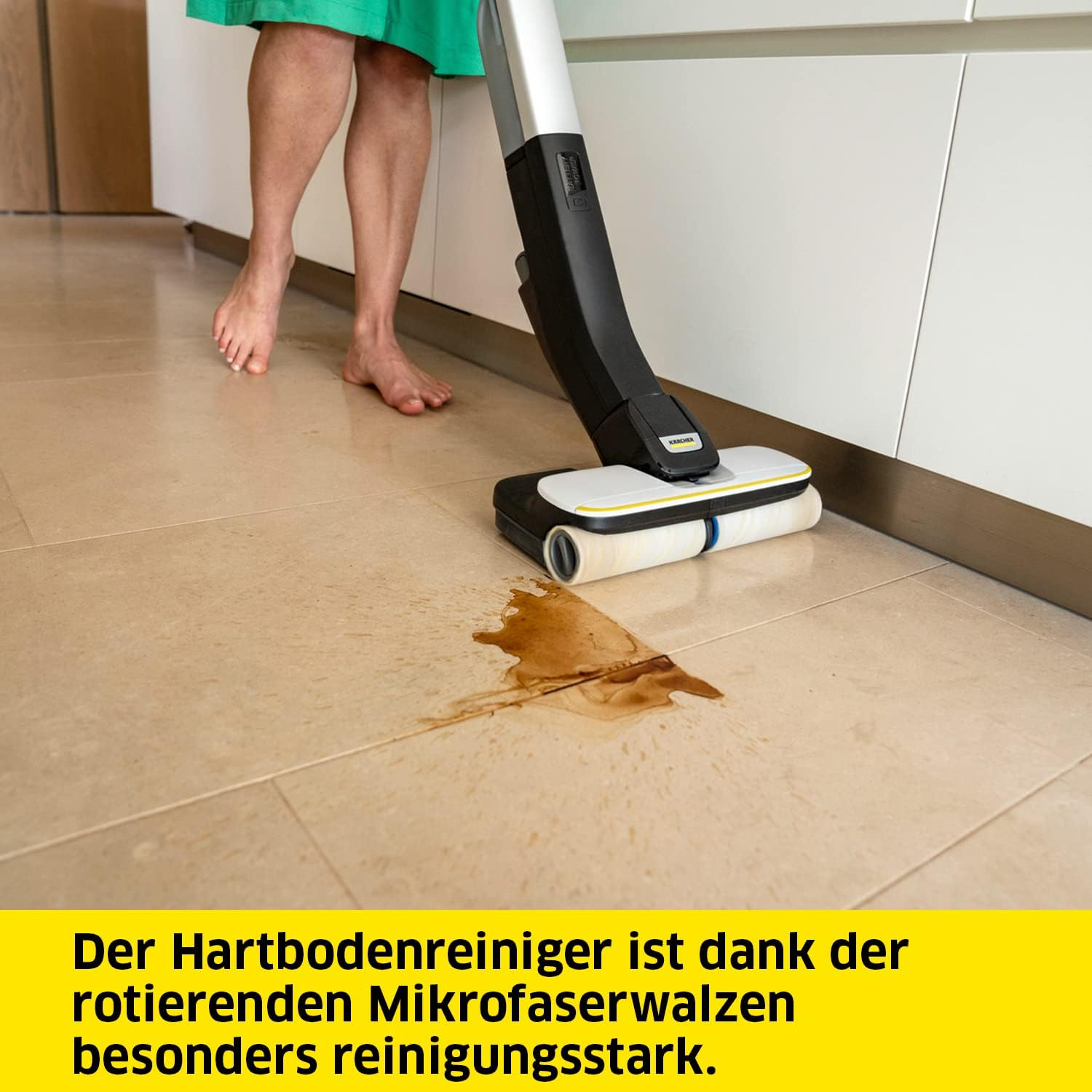 Osoba czyszcząca kafelki za pomocą myjki do podłóg. Na podłodze brązowa plama. Białe szafki.