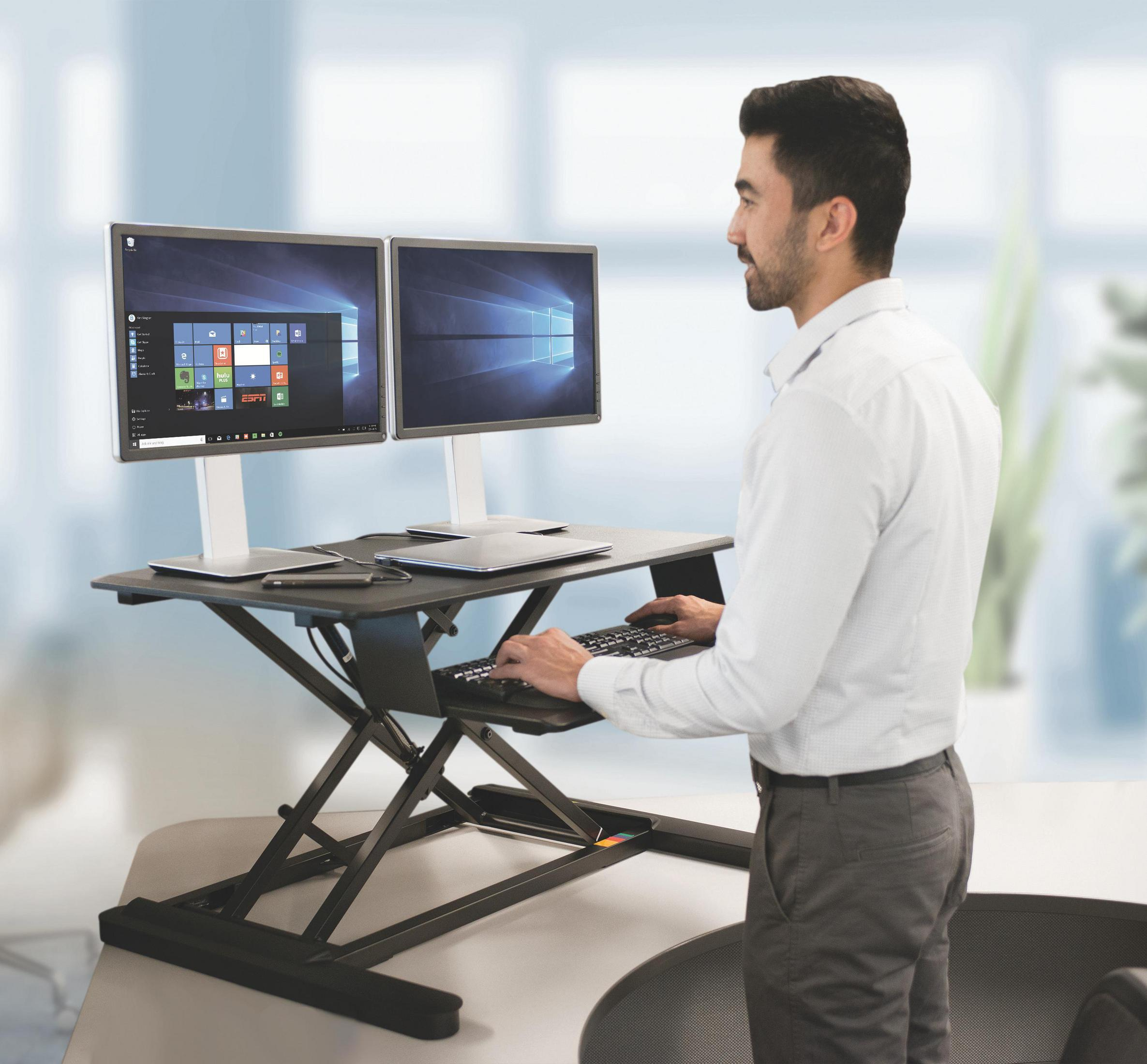 Mann, der an einem Stehpult mit zwei Monitoren, Tastatur und Stift arbeitet. Weißer Hintergrund.