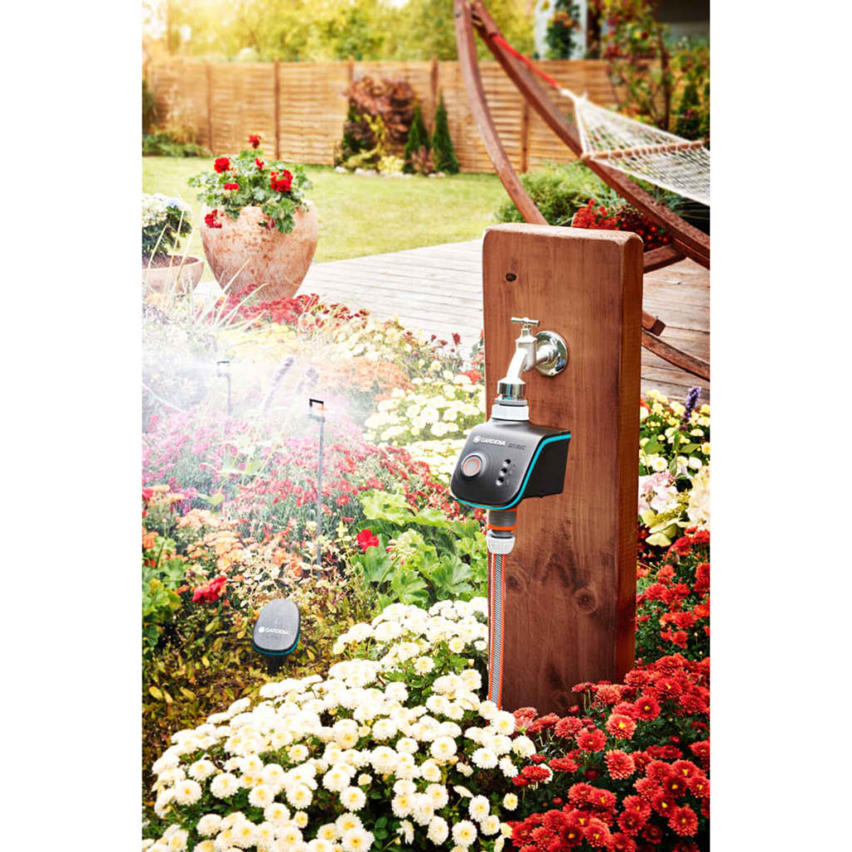 Una scena di giardino con un sistema di irrigazione. Un palo di legno sostiene un rubinetto e un'unità di controllo. Fiori ed erba.