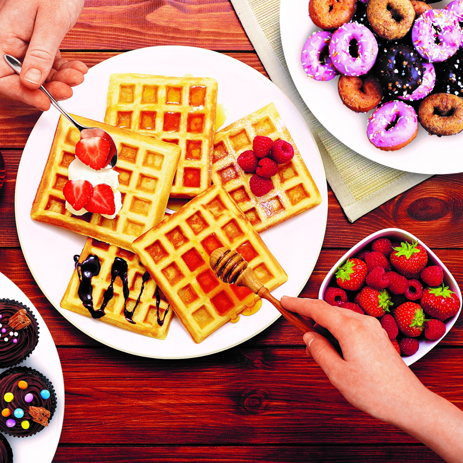 Gofry z dodatkami, pączki, babeczki i dłonie serwujące. Drewniany stół.