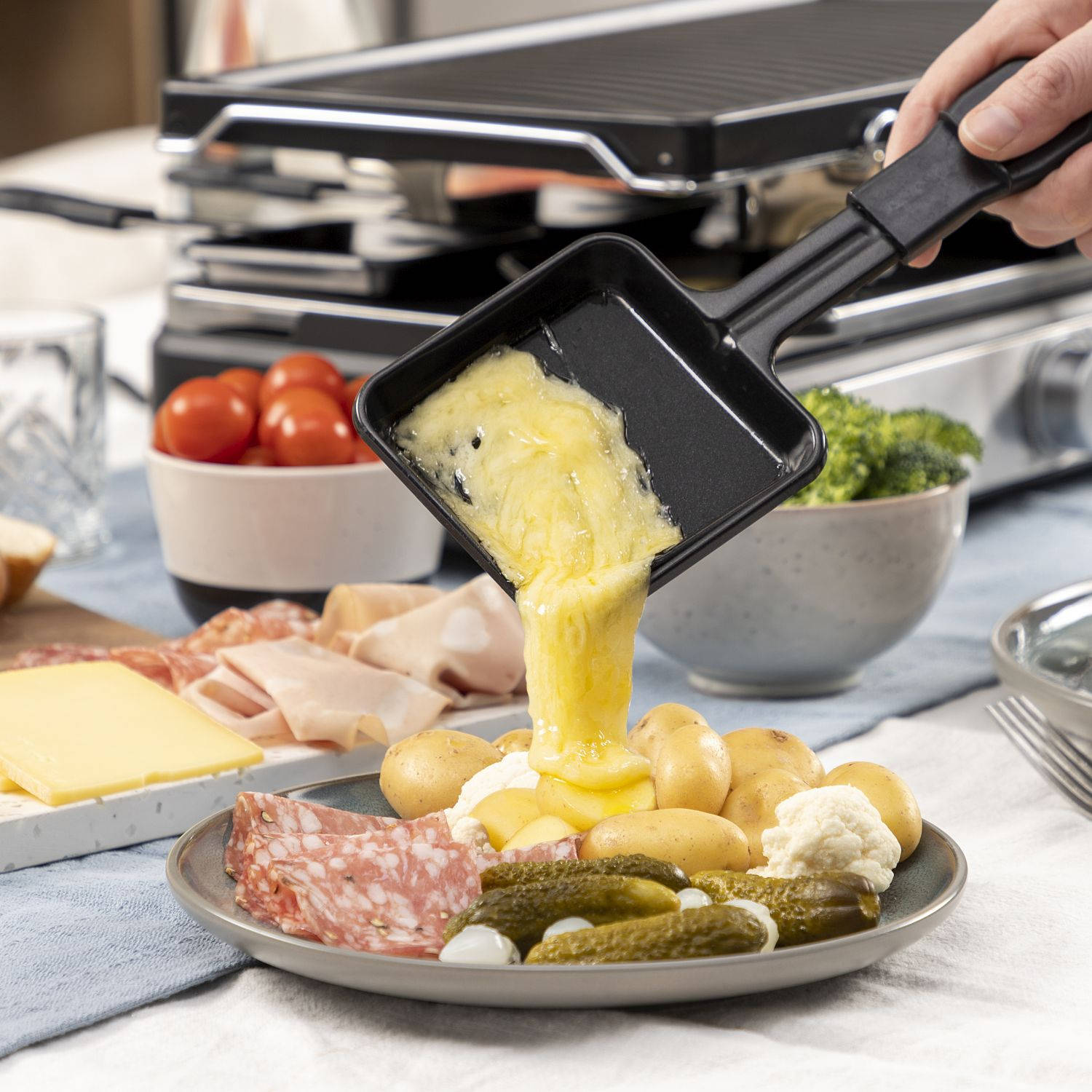 Eten wordt uit een raclettepan op een bord gegoten met aardappelen, augurken en salami. Witte achtergrond.