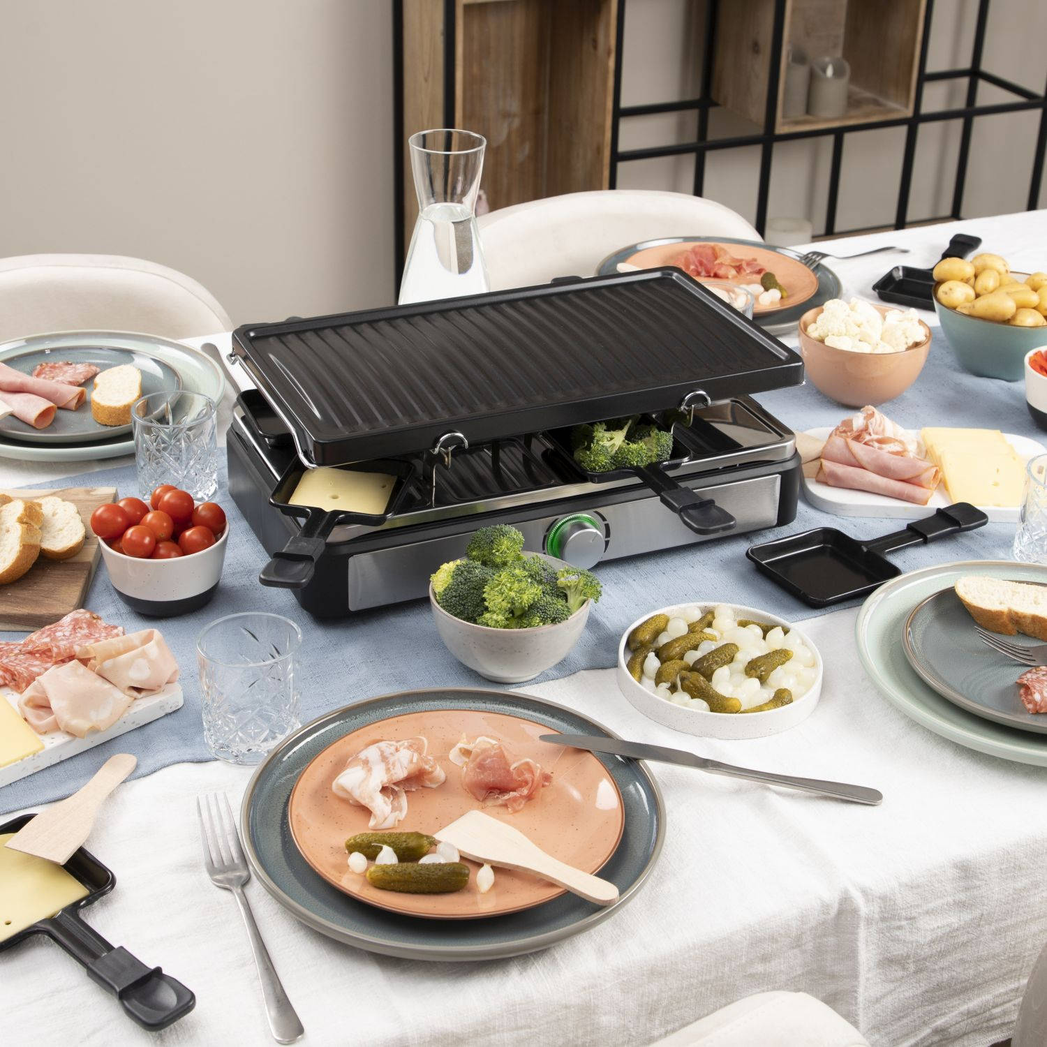 Een eettafel met een raclette grill, etenswaren zoals kaas, vlees en groenten op de tafel.