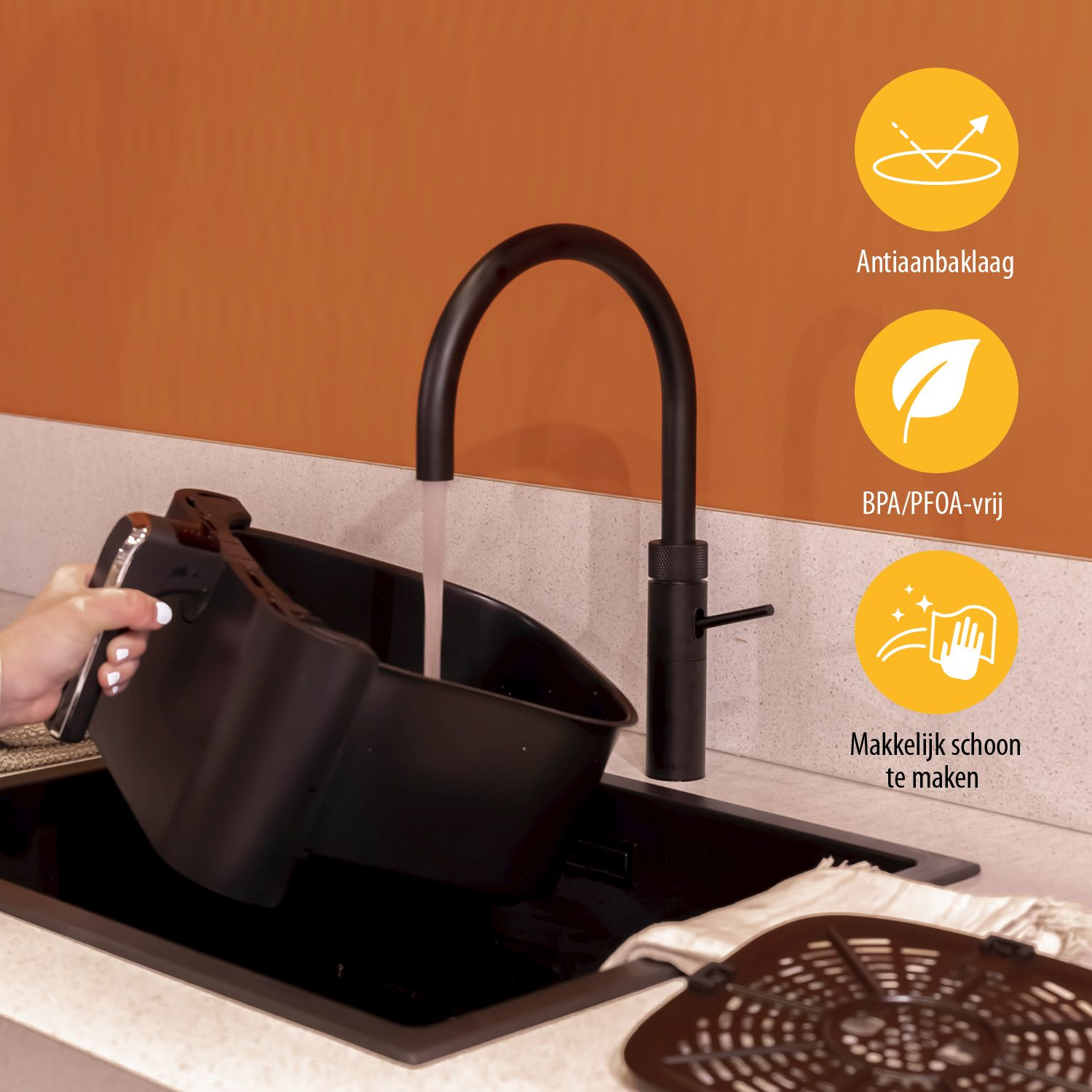 Osoba trzymająca czarny kosz do frytkownicy powietrznej, w pobliżu czarnej baterii i zlewu. Biały blat i pomarańczowa ściana.