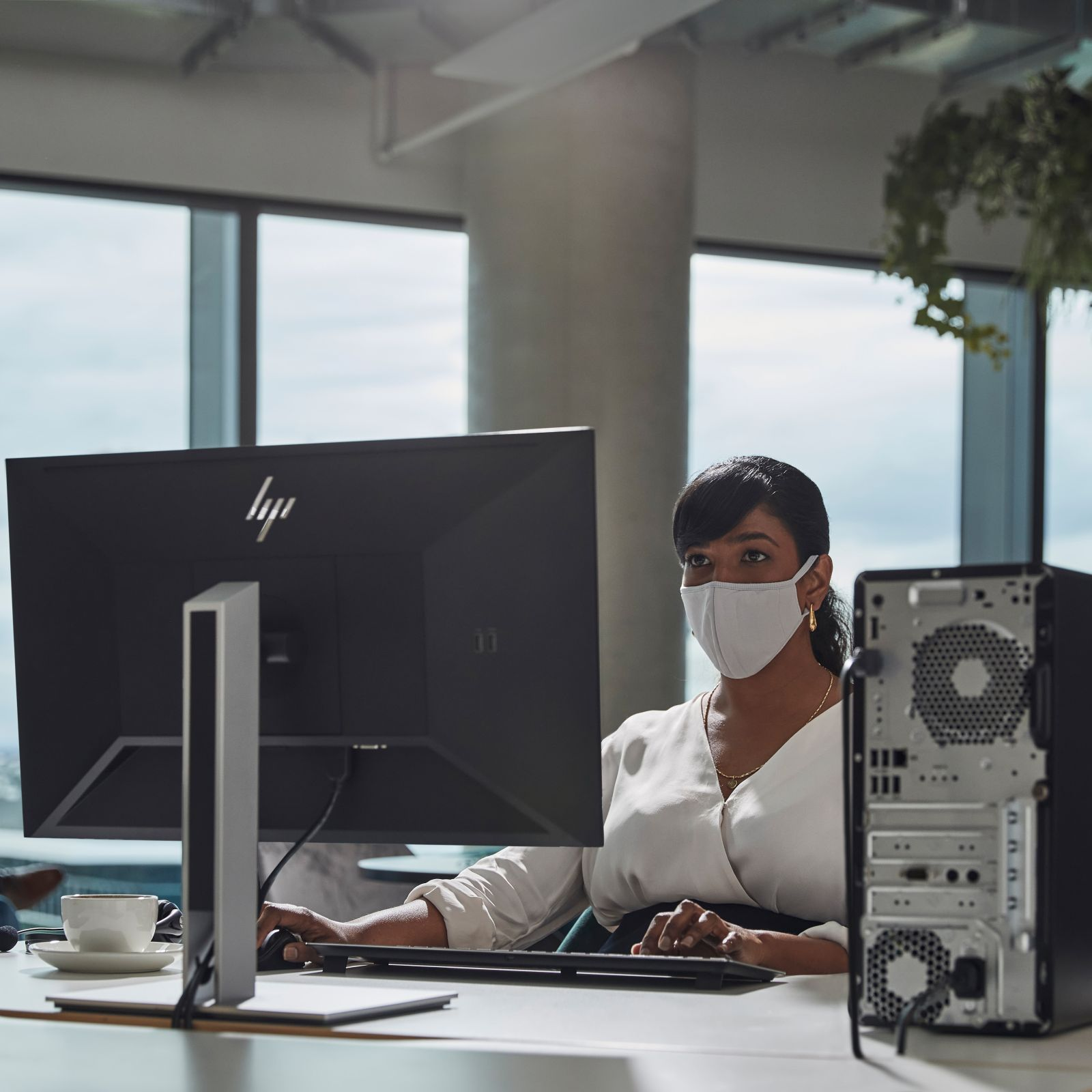 Kobieta w masce przy komputerze w nowoczesnym biurze. Widoczny monitor i tył wieży.