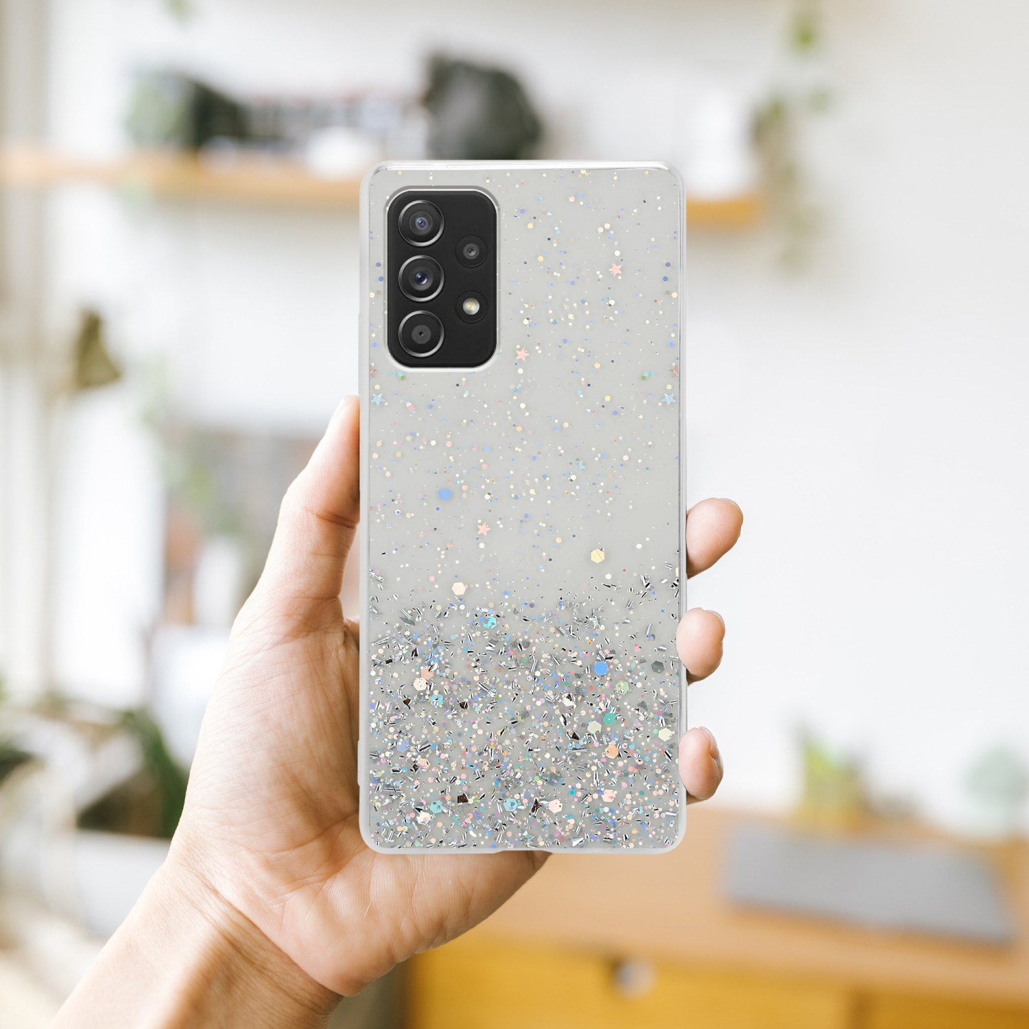 Una mano che tiene un telefono in una custodia trasparente con glitter e stelle. È visibile uno sfondo sfocato.