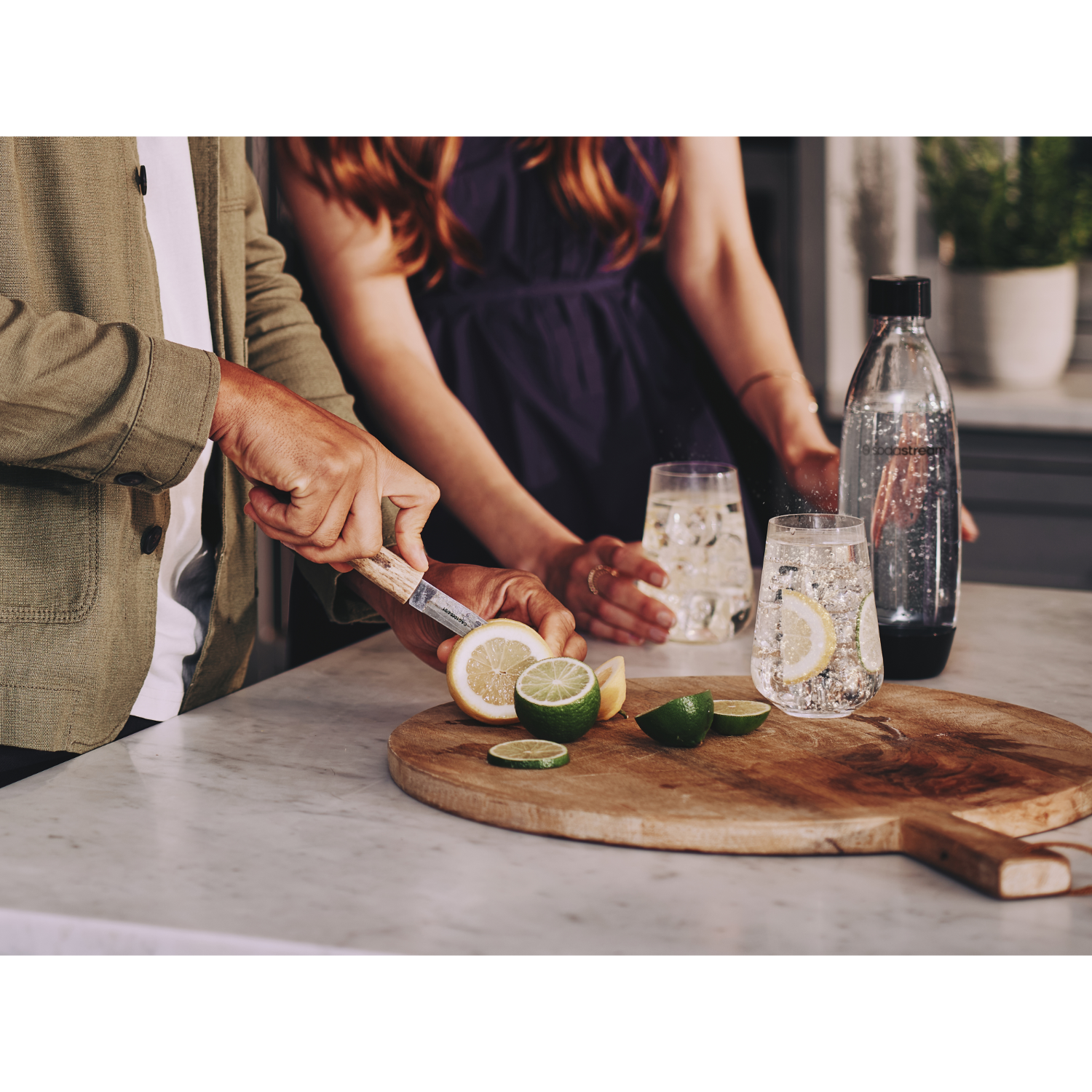 Osoba kroi cytrynę. Dwie szklanki z wodą, plasterki cytryny i limonki. Butelka wody sodowej po prawej.