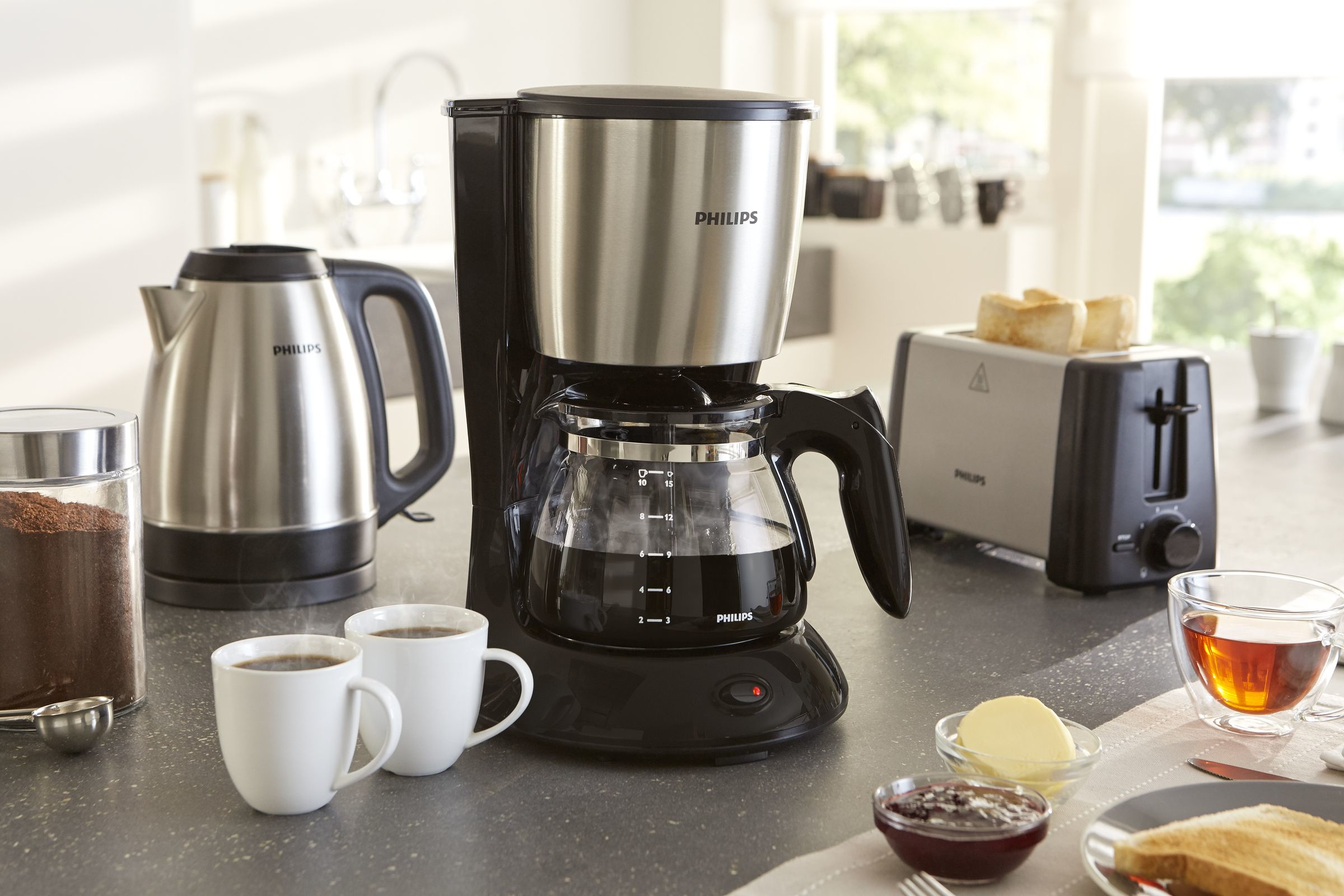 Encimera de cocina con cafetera, hervidor, tostadora y desayuno. Hay dos tazas de café.