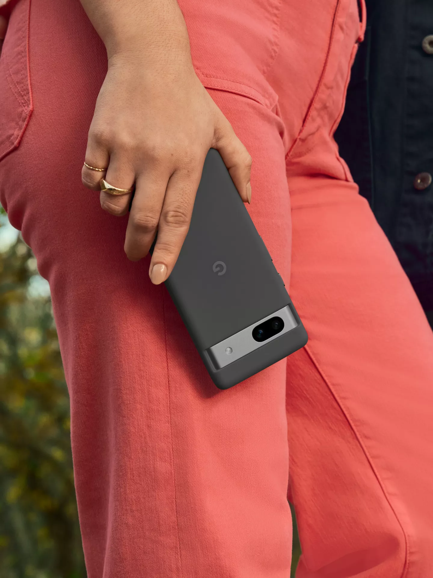 Una mano che tiene un telefono grigio scuro Google Pixel 7a, con anelli d'oro, pantaloni rossi.