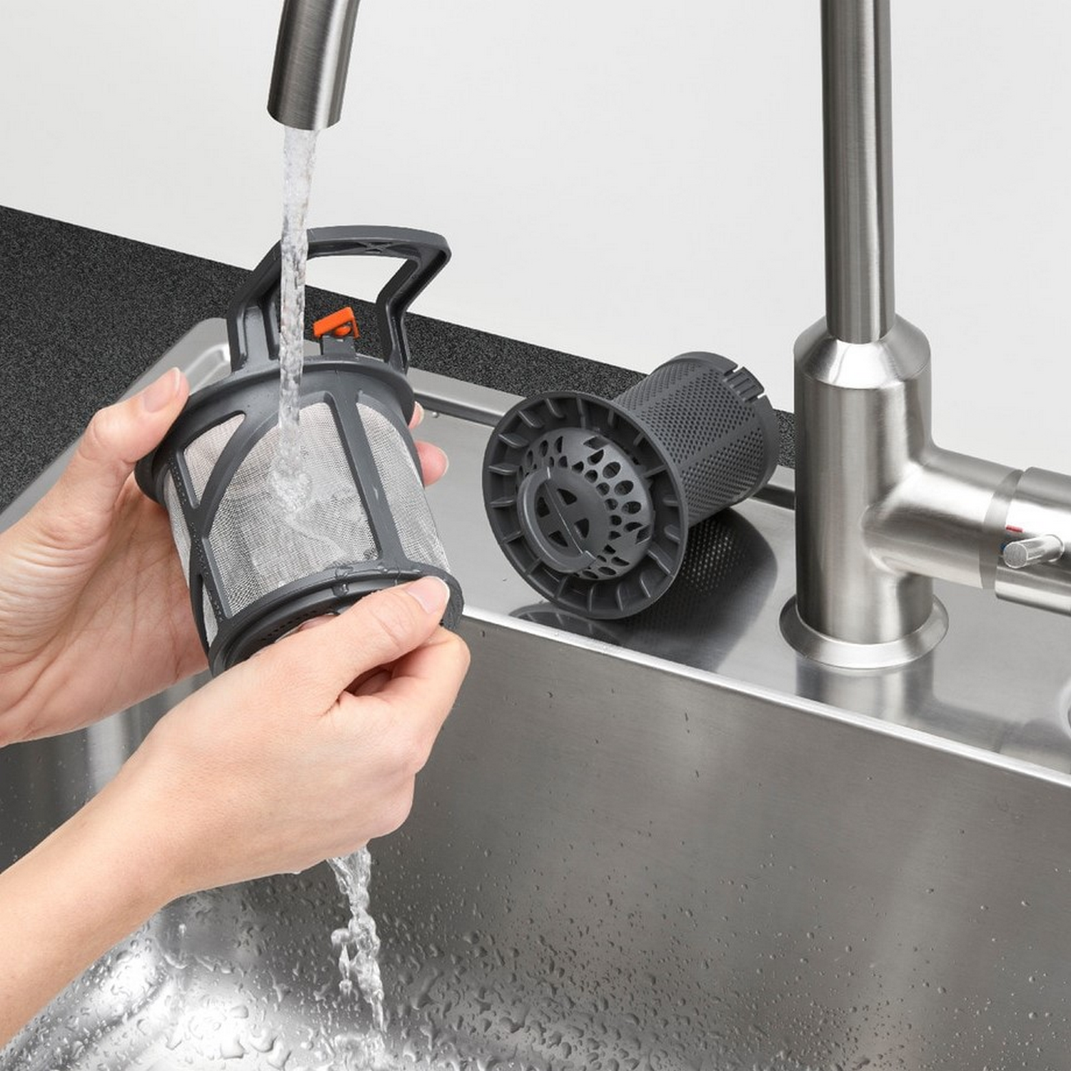 Ręce trzymają filtr zmywarki pod bieżącą wodą. Drugi filtr leży obok zlewu.