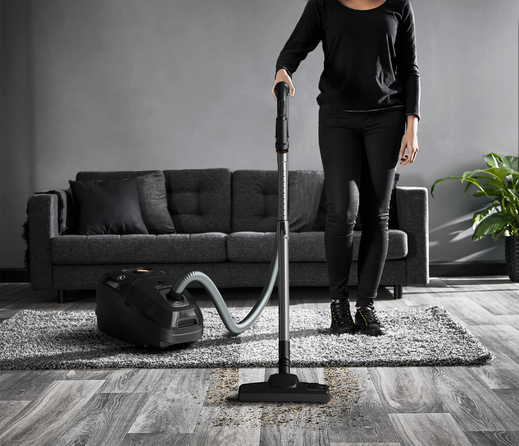 Osoba odkurzająca pokój. Odkurzacz jest czarny, pokój ma drewniane podłogi i sofę.