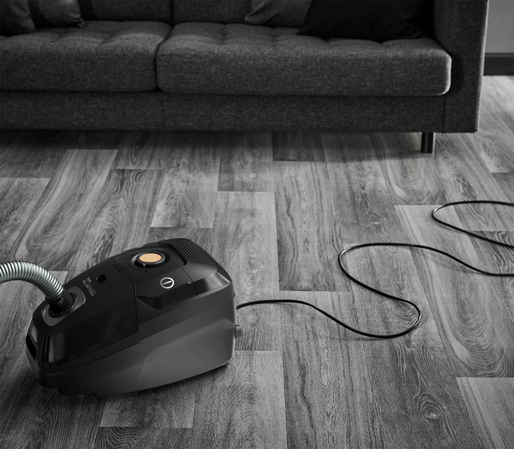 Czarny odkurzacz na drewnianej podłodze obok szarej sofy.