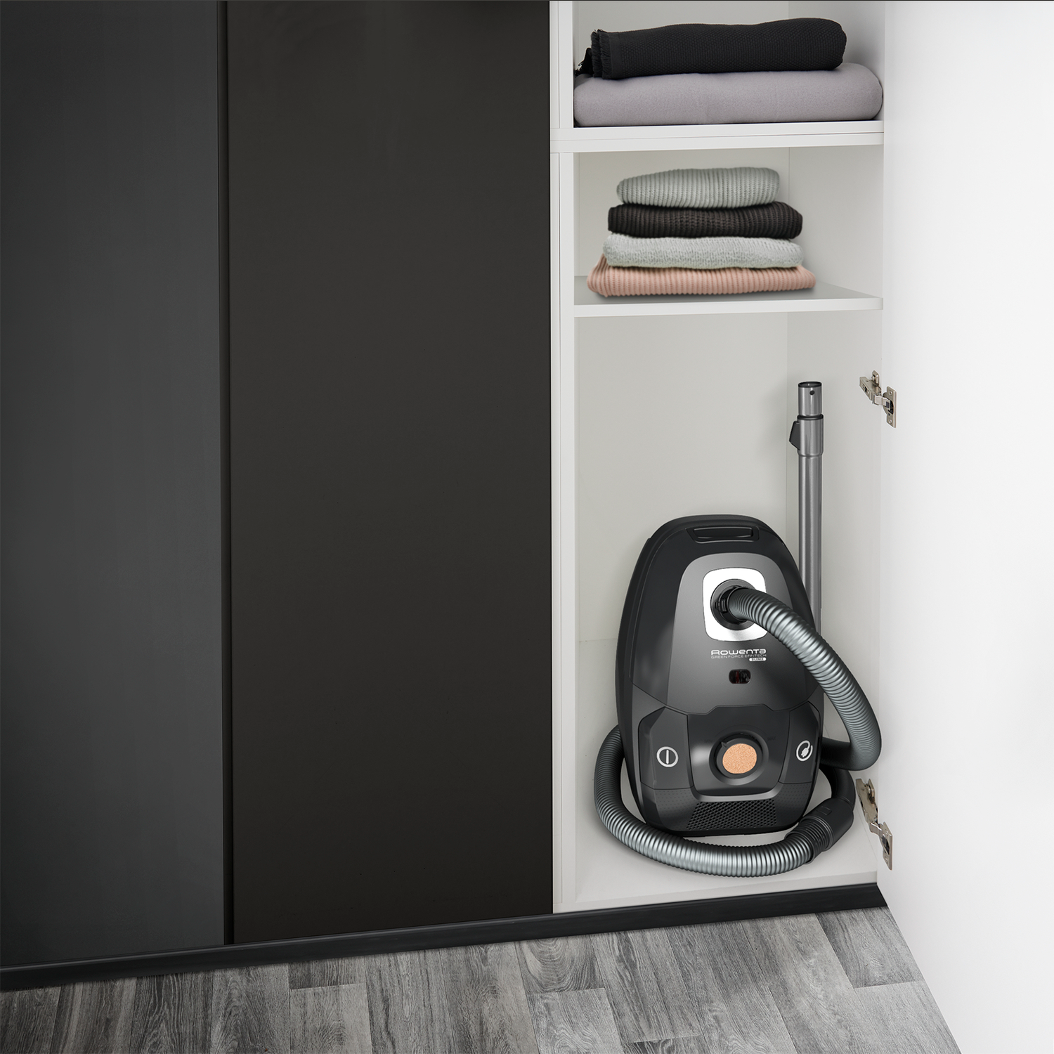 Otwarta szafa z ciemnoszarym odkurzaczem, srebrnym wężem i ułożoną złożoną odzieżą na półkach.
