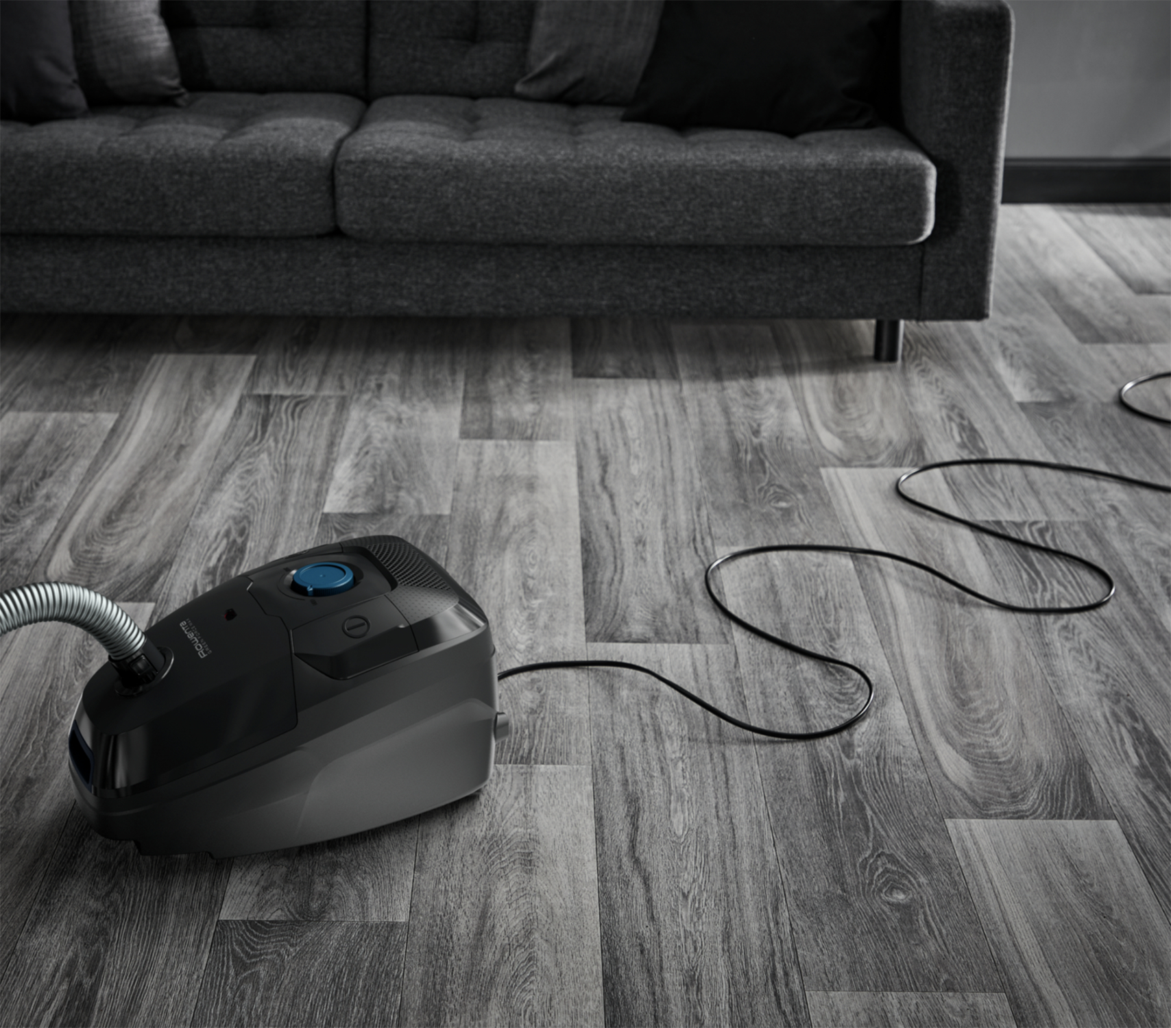 Czarny odkurzacz na drewnianej podłodze. Szara sofa w tle.