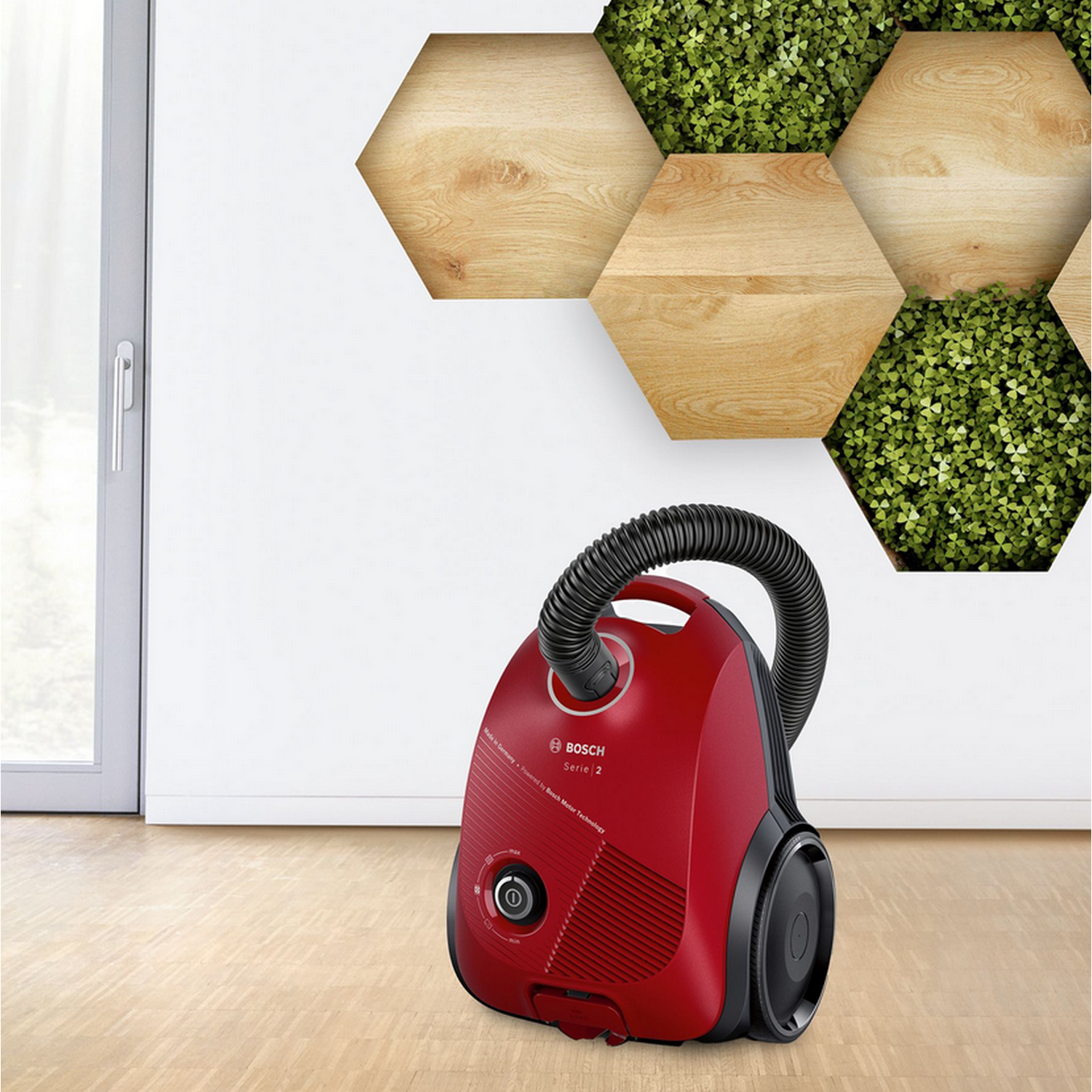 Czerwony odkurzacz Bosch stoi na drewnianej podłodze obok ściany z sześciokątnym wzorem.