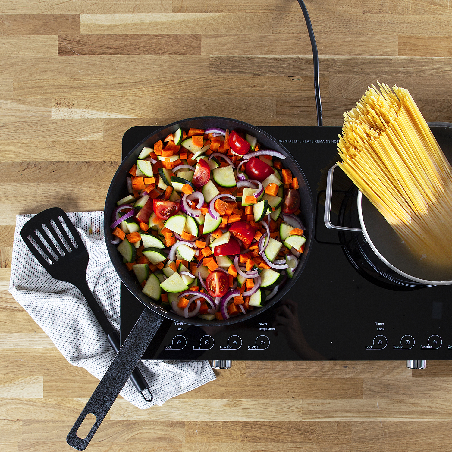 Draufsicht auf eine Pfanne mit Gemüse, Spaghetti und einem Pfannenwender auf einer Holzoberfläche.