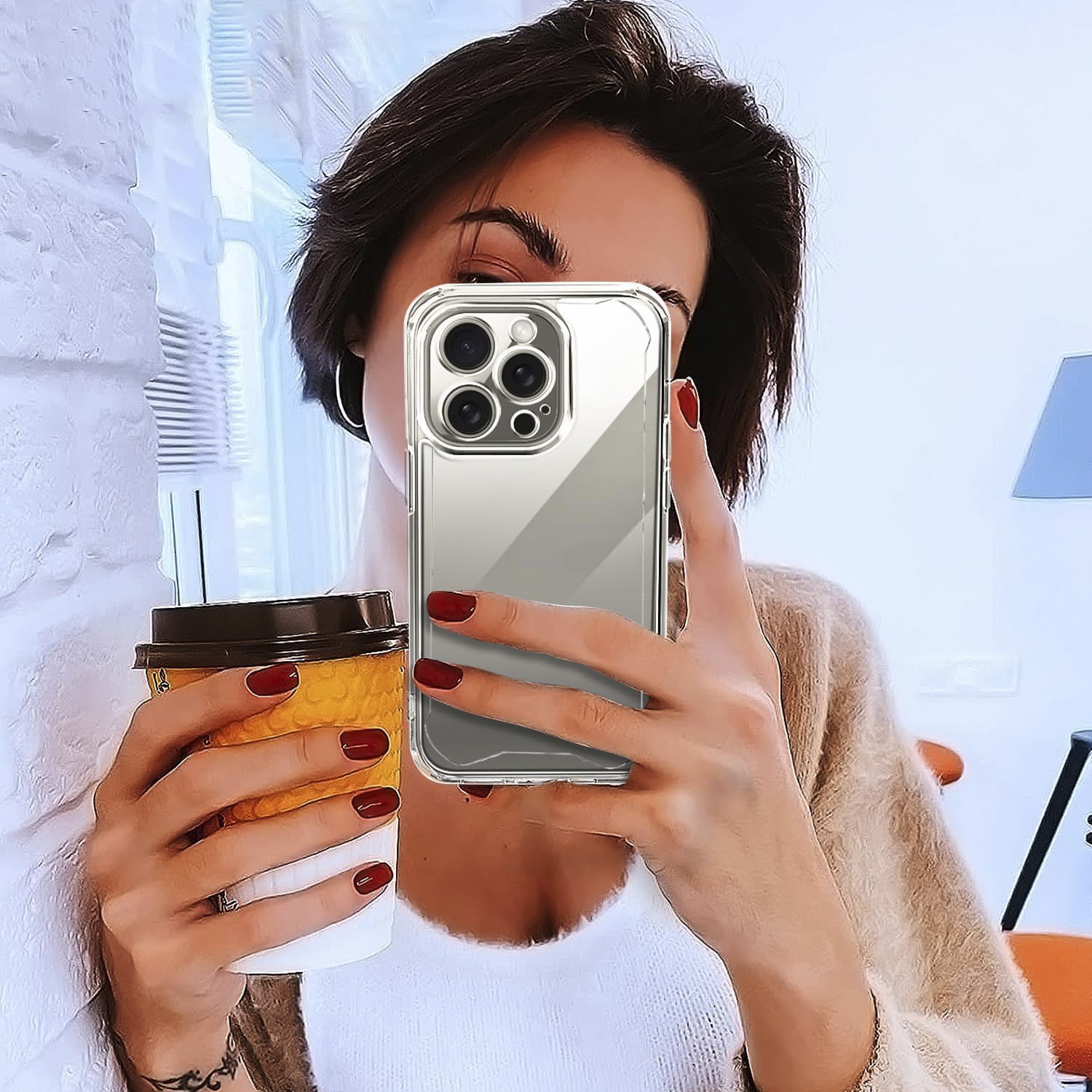 Donna che tiene in mano un telefono e una tazza di caffè, con unghie rosse. Muro di mattoni bianchi.