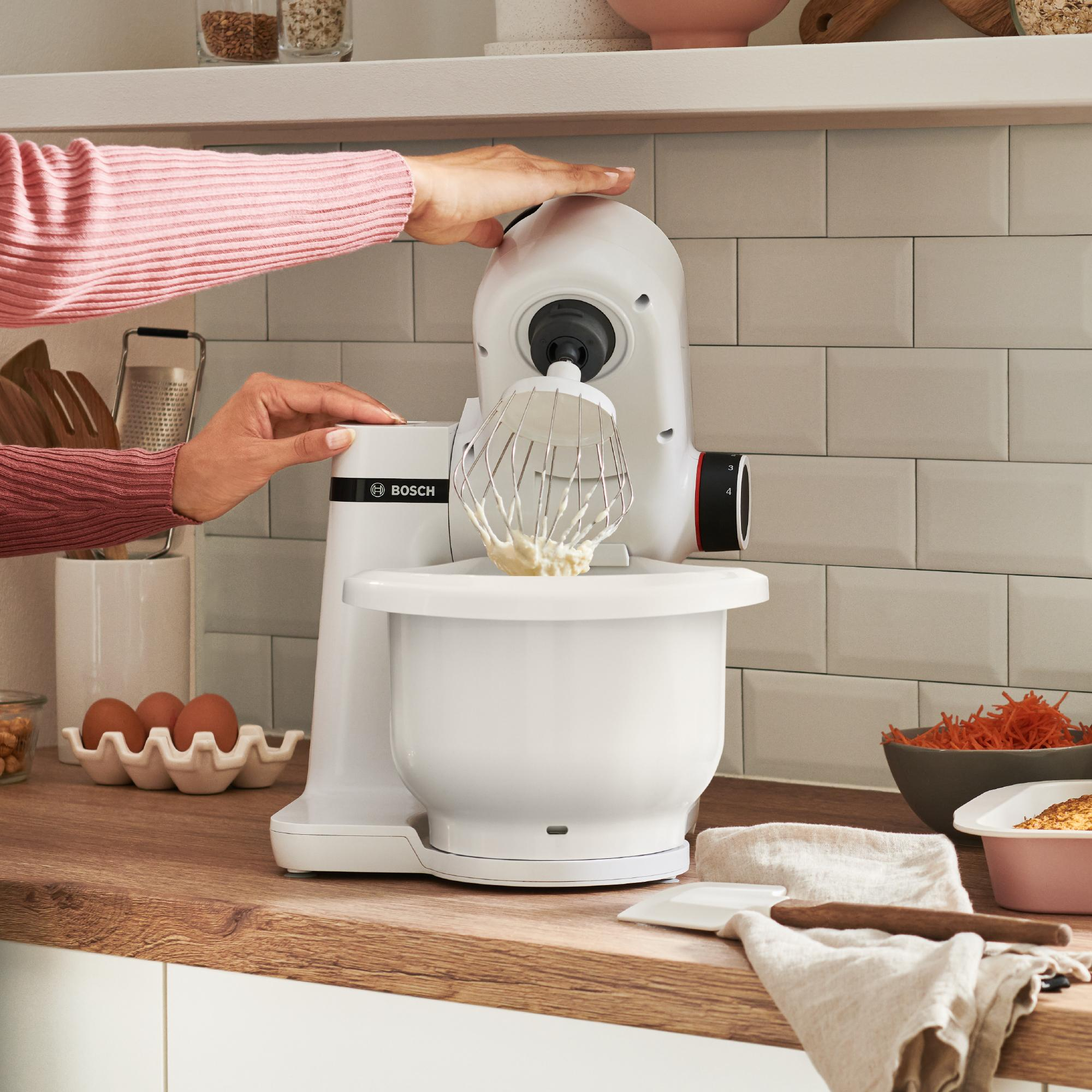 Ein weißer Küchenmixer in Betrieb mit einer Schüssel Teig und Zutaten in der Nähe.