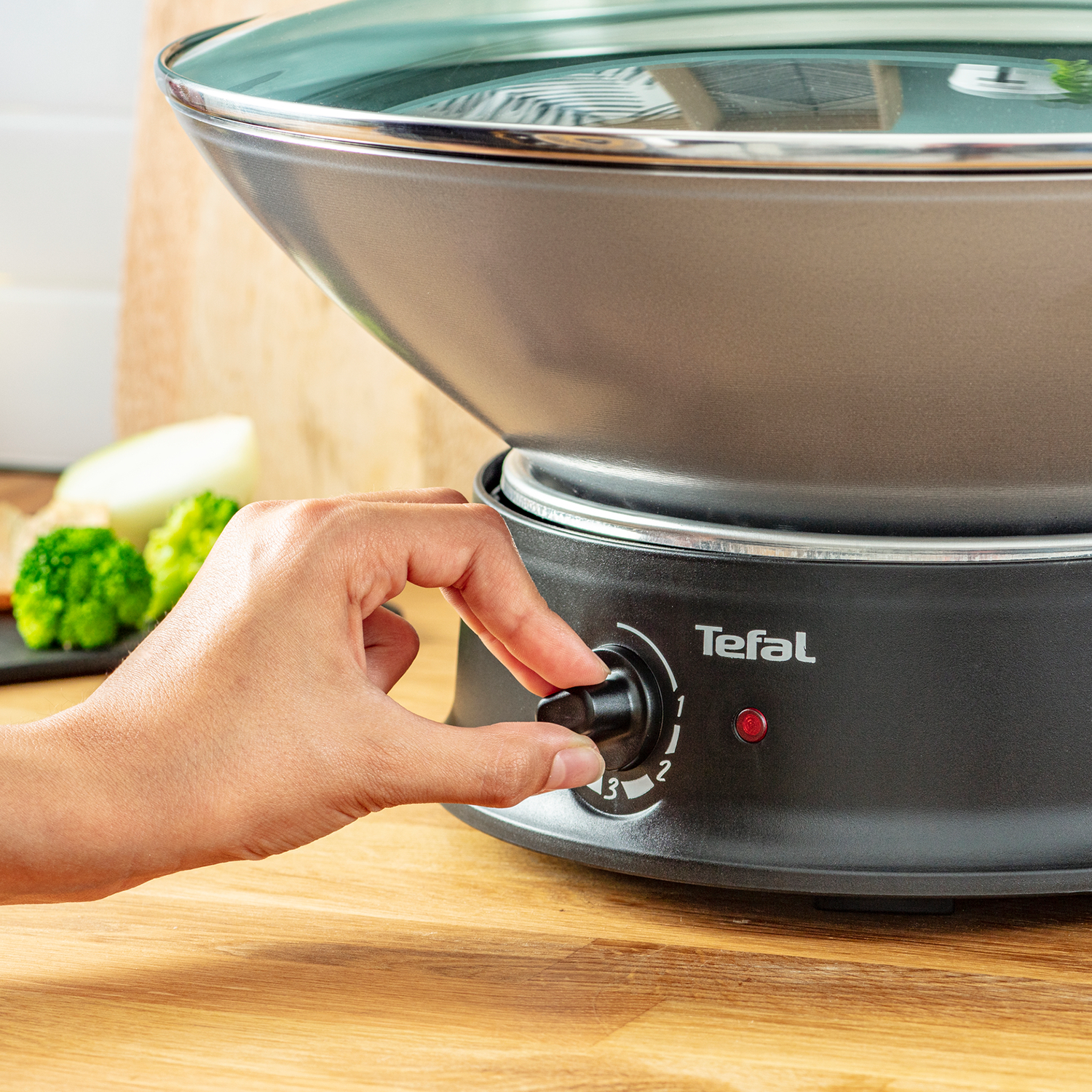Een hand draait aan de knop van een zwarte Tefal wok op een houten oppervlak, rood licht brandt.
