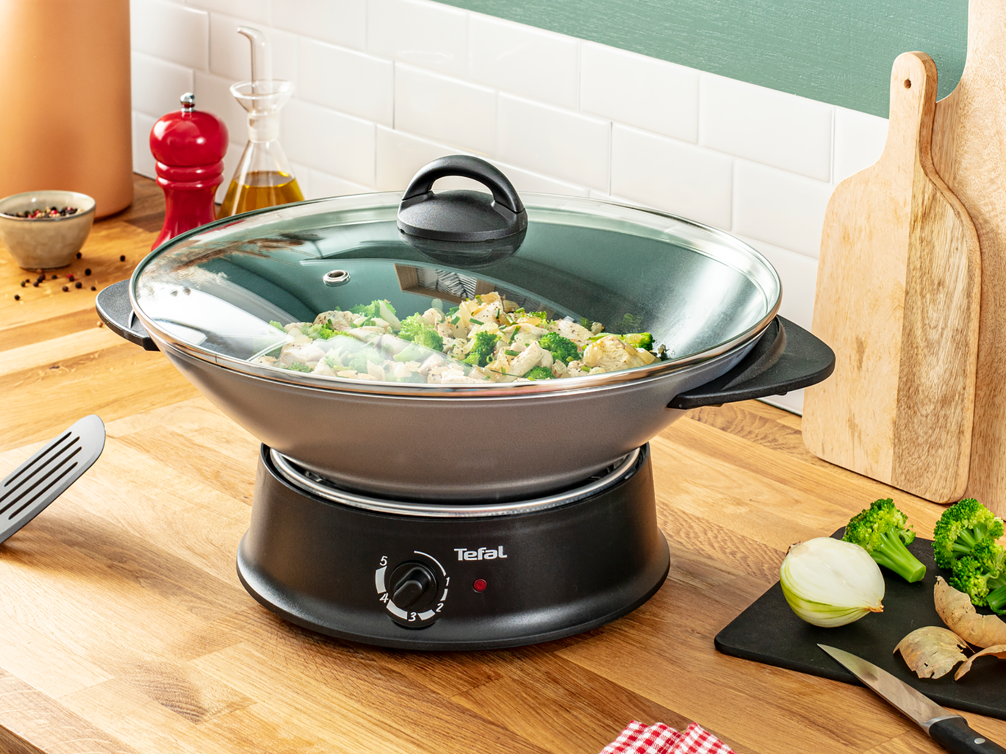 Een wok met eten staat op een zwarte elektrische basis. De deksel is helder. Er is een houten oppervlak.