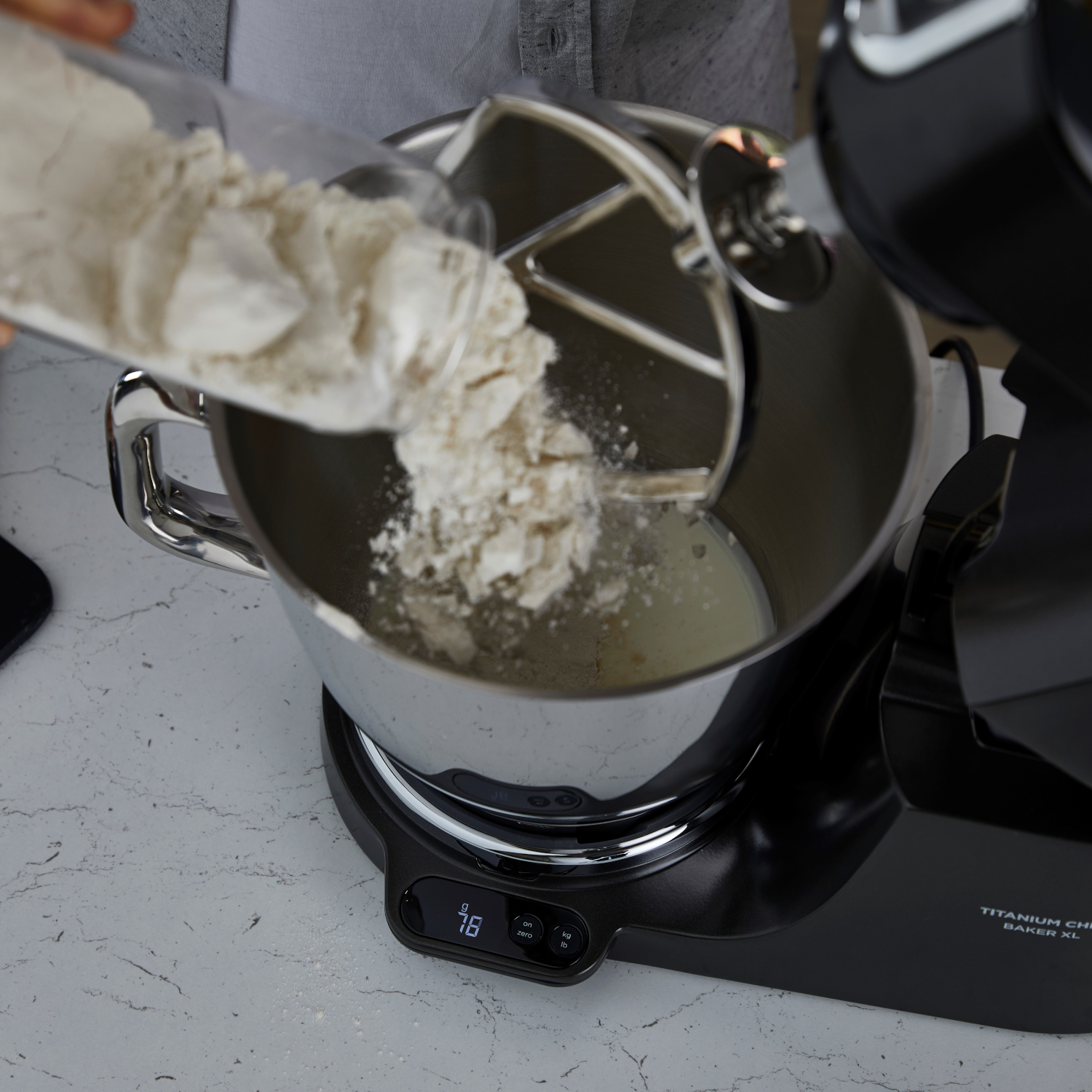 Farina versata in una ciotola. L'impastatrice è nera, con ciotola e braccio in acciaio inox.