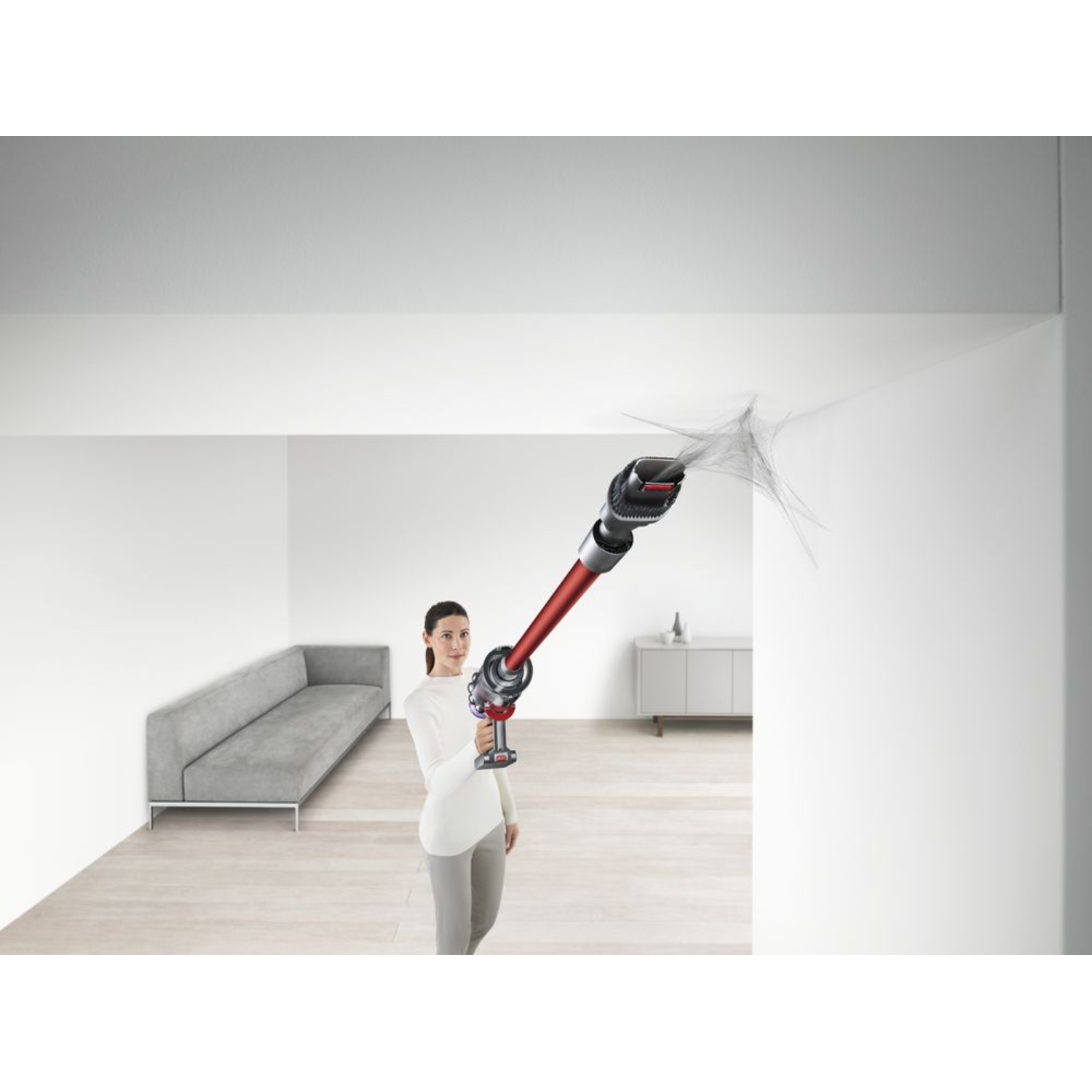 Kobieta odkurza ścianę. Odkurzacz w ręku, sprzątanie. Biały pokój, szara kanapa.