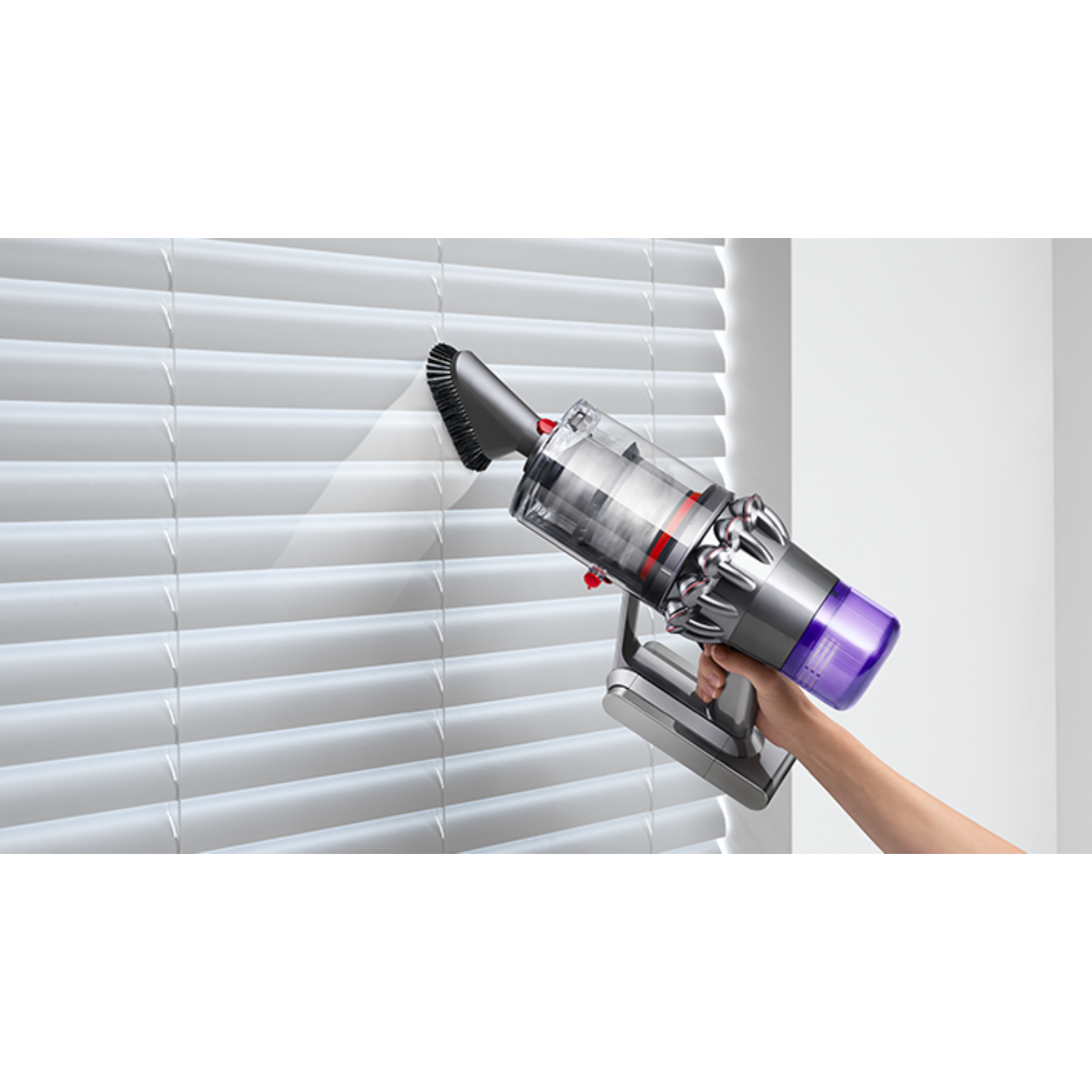 Ręka trzyma przystawkę do odkurzacza czyszczącą białe żaluzje. Odkurzacz jest szary i fioletowy.
