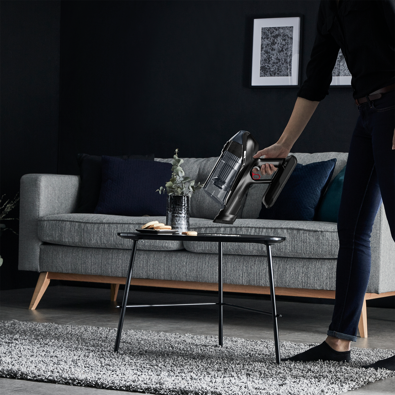 Eine Person hält einen schwarzen, kabellosen Staubsauger auf einem grauen Sofa, schwarzer Tisch.