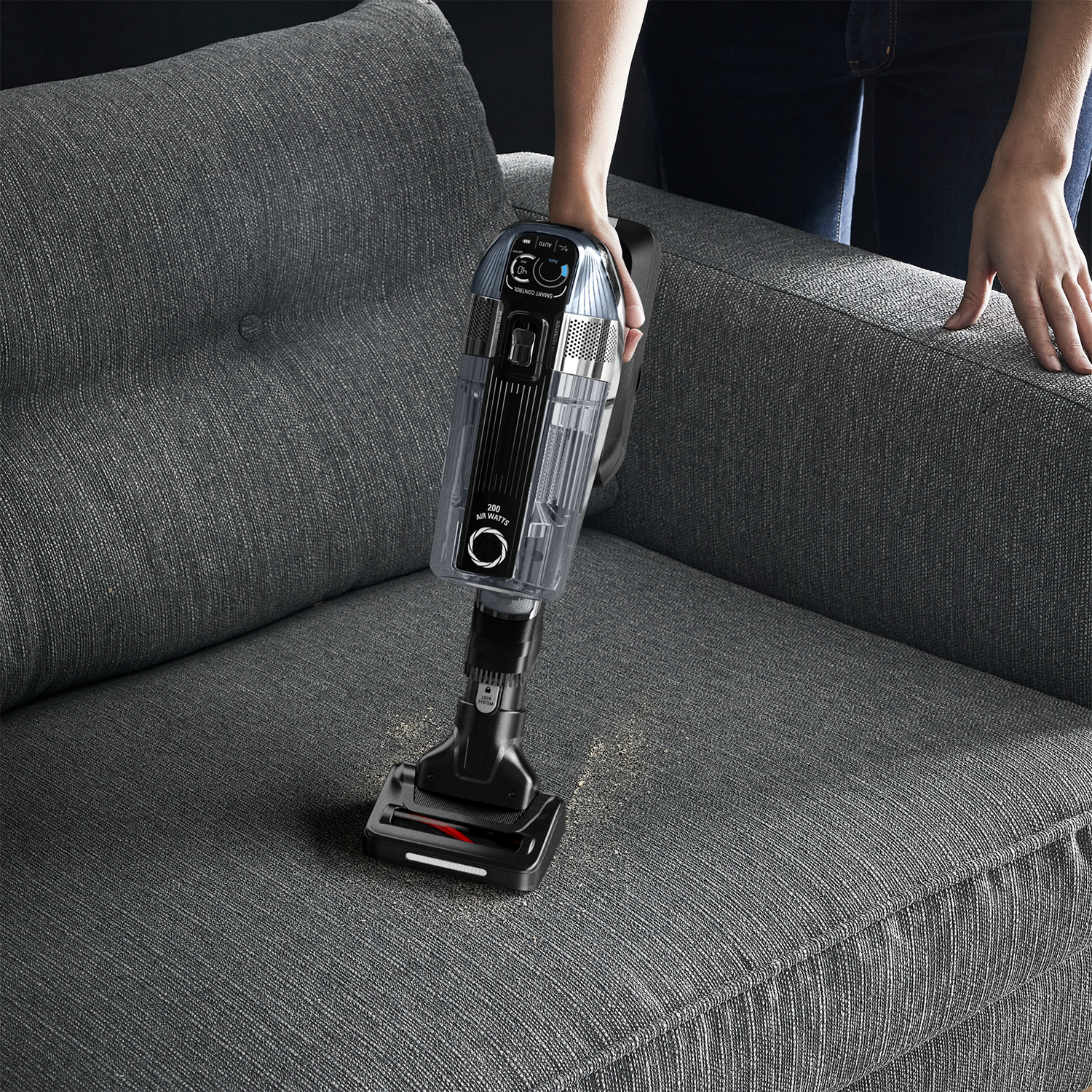 Eine Person saugt ein graues Sofa mit einem schwarz-transparenten Staubsauger ab. Staub wird aufgesaugt.