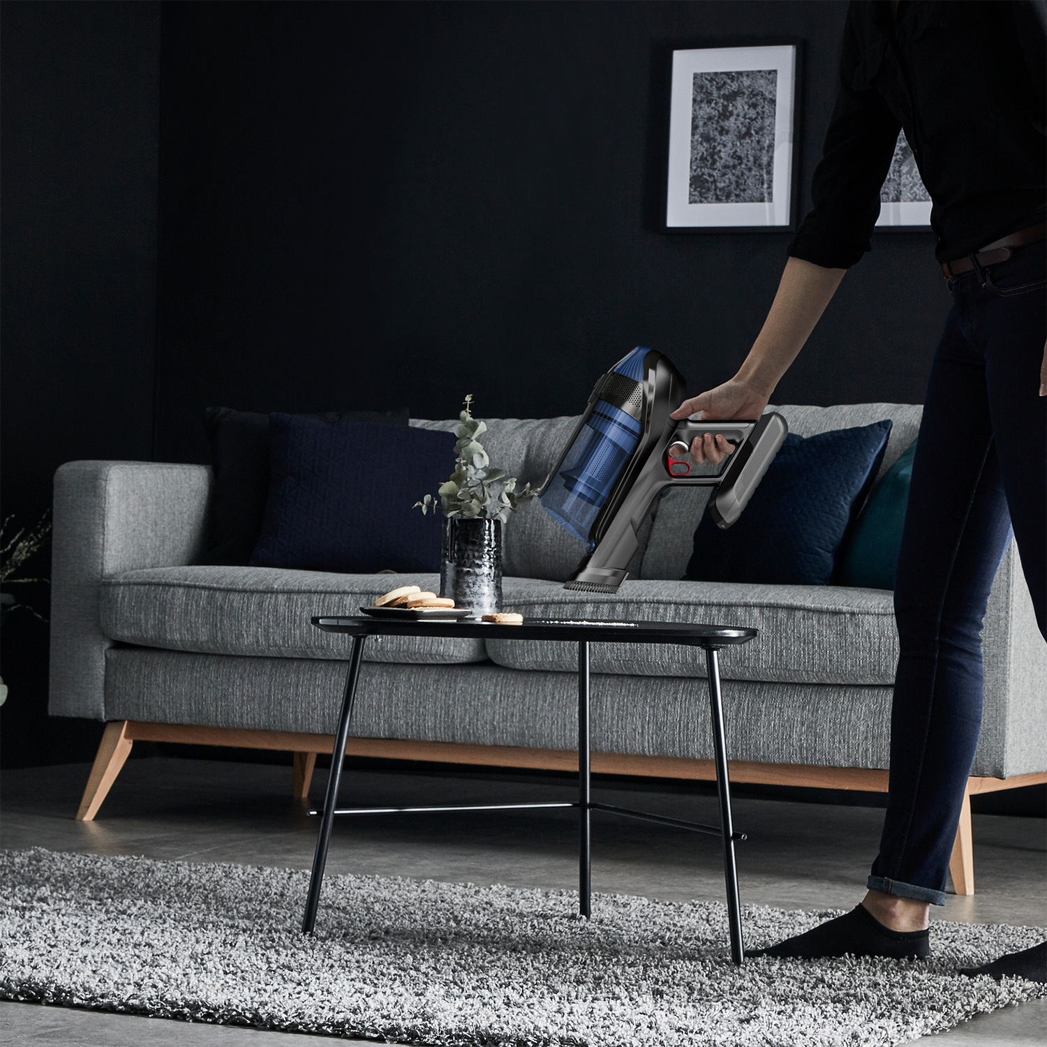 Eine Person, die einen grauen Teppich mit einem kabellosen Staubsauger saugt. Graues Sofa, schwarzer Tisch und Kunst an der Wand.