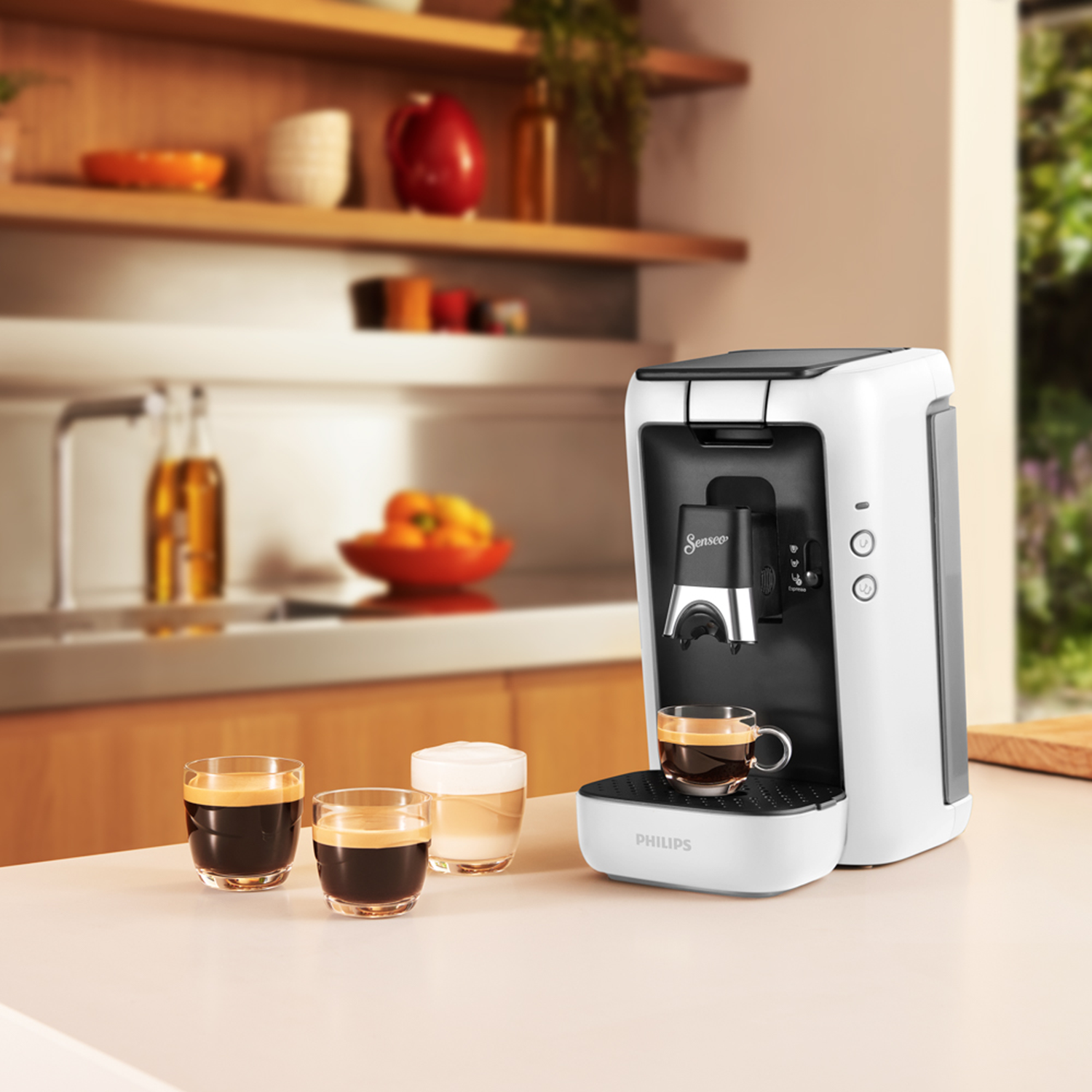 Biały ekspres do kawy z dwiema filiżankami kawy, trzecia z mlekiem, na stole, rozmyte tło.