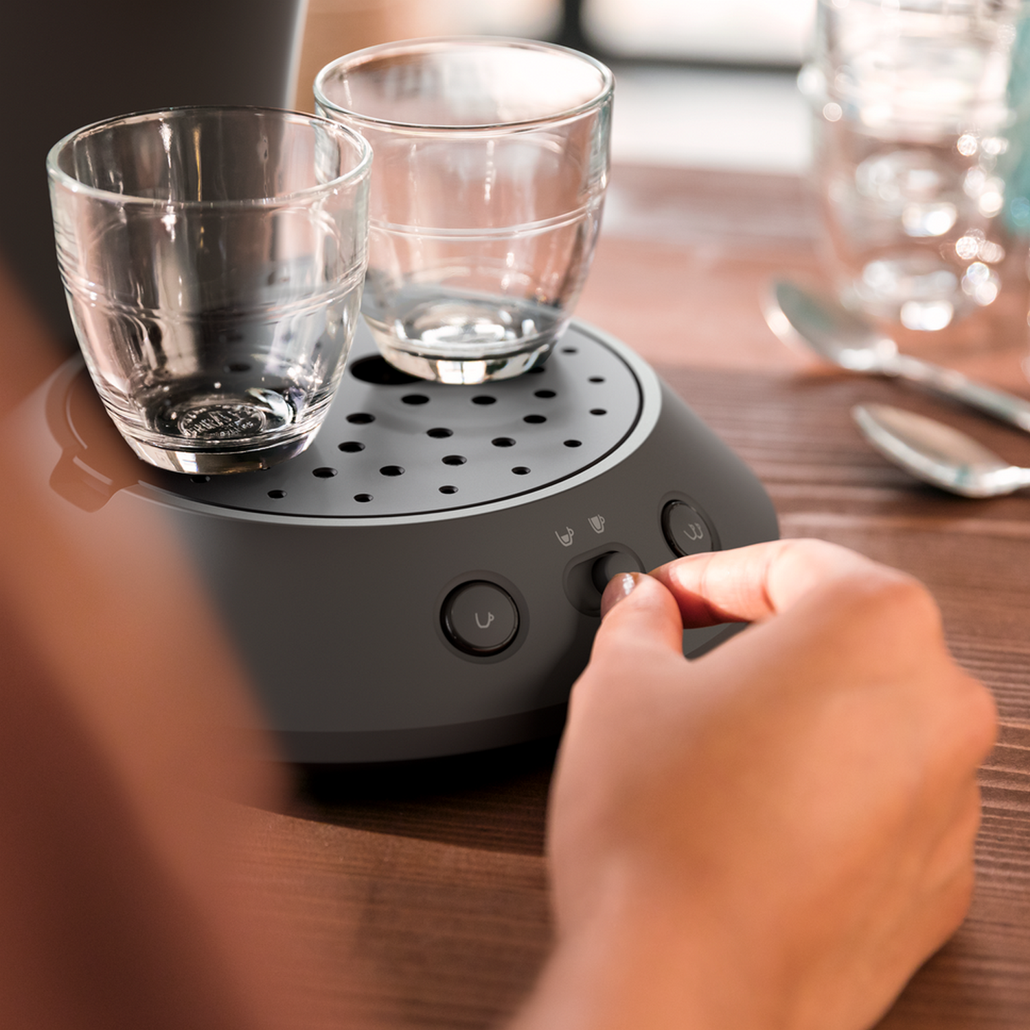 Ręka obraca pokrętło w ekspresie do kawy. Na górze dwa kieliszki. Łyżki i szklanka wody w tle.