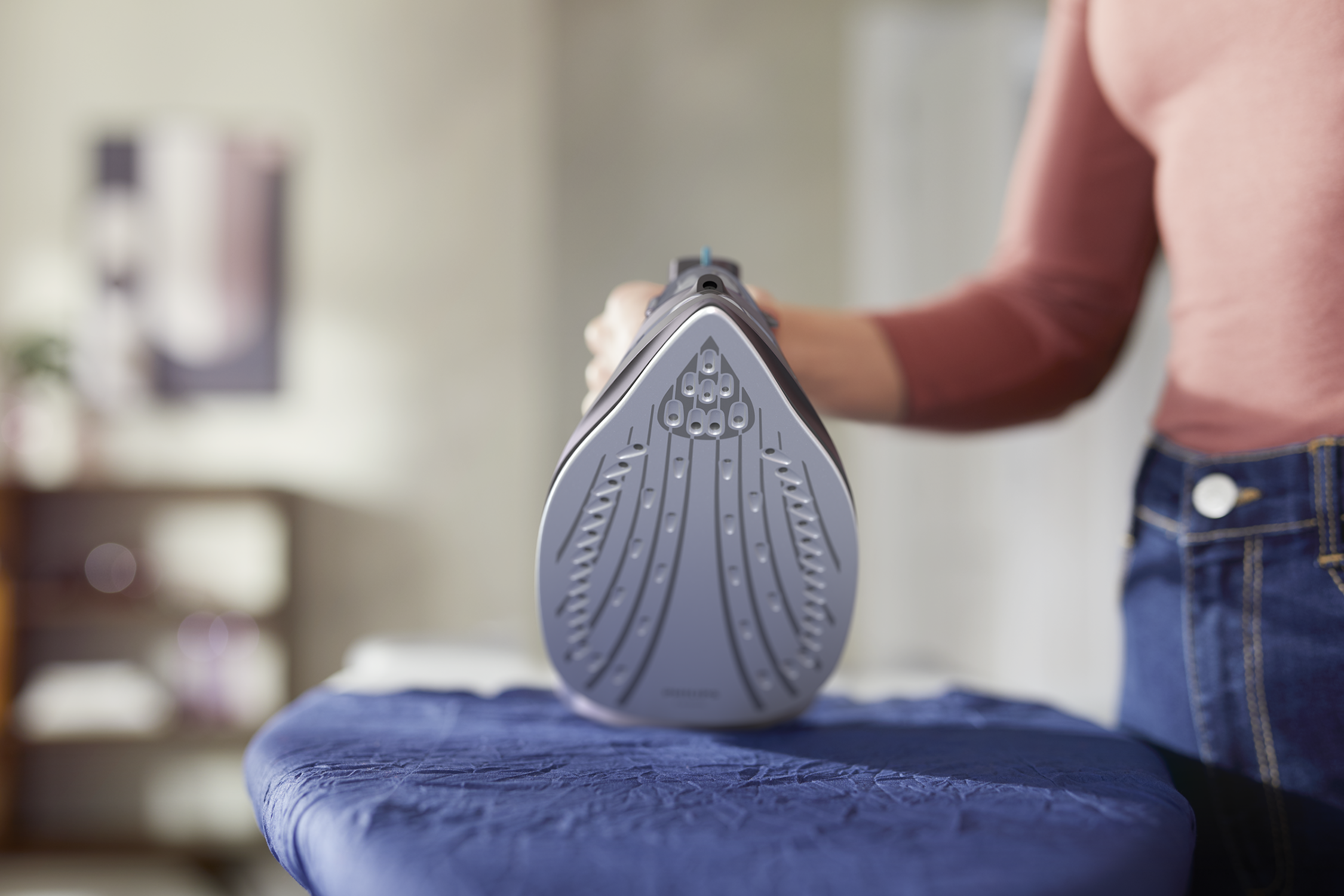 Osoba trzymająca szare żelazko nad niebieską deską do prasowania, w pokoju.