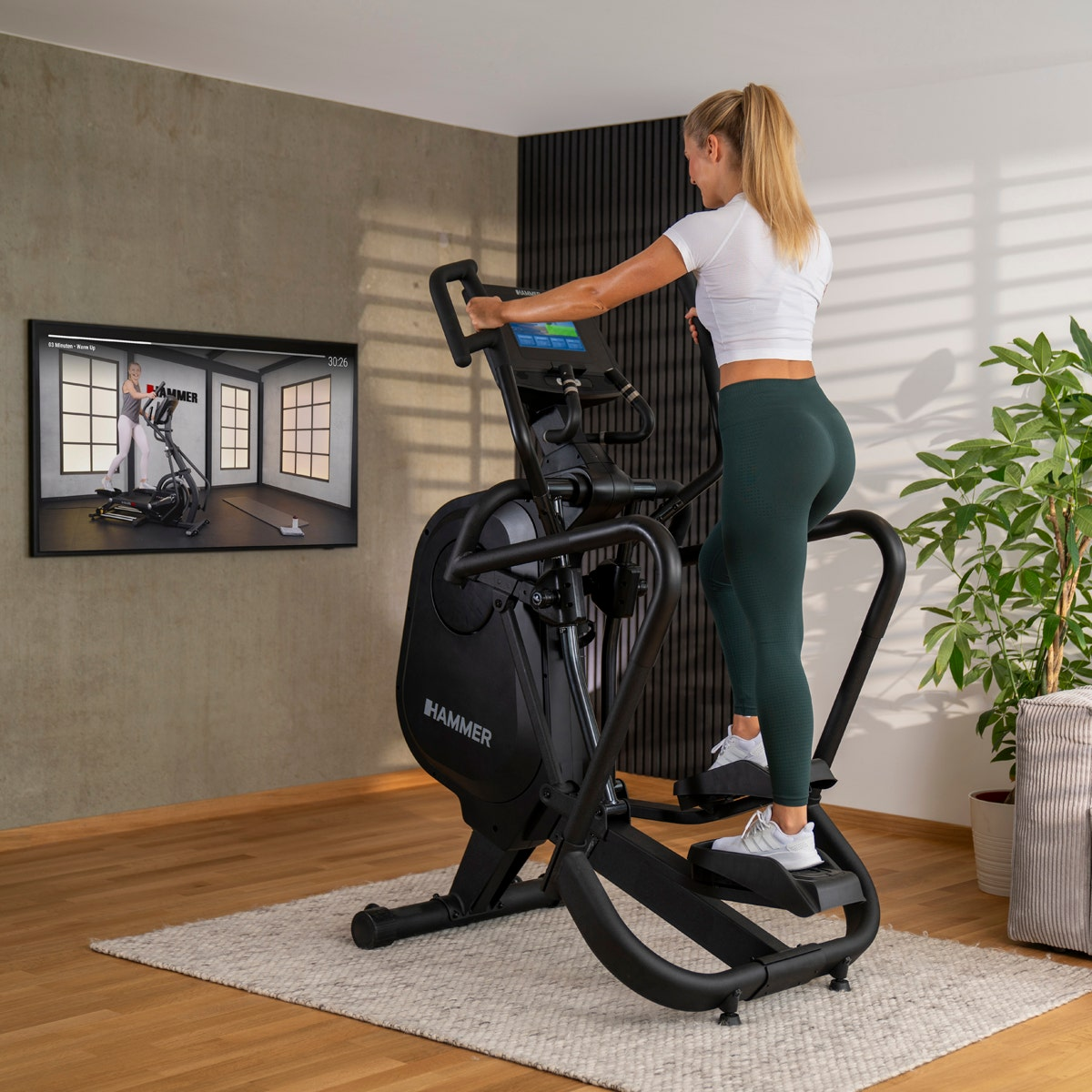 Frau auf Crosstrainer, schaut TV. Schwarze Maschine, grüne Leggings, hell.