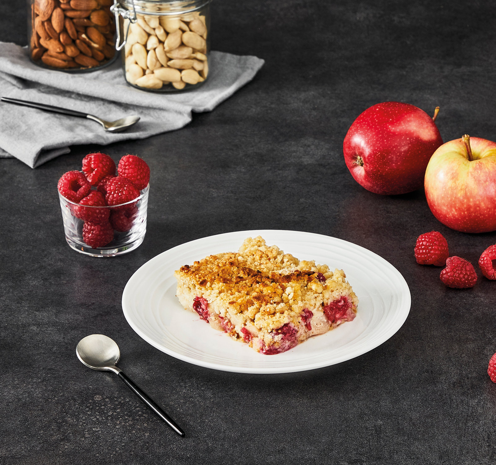 A plate of crumble with raspberries and apples, with jars of almonds and nuts.
