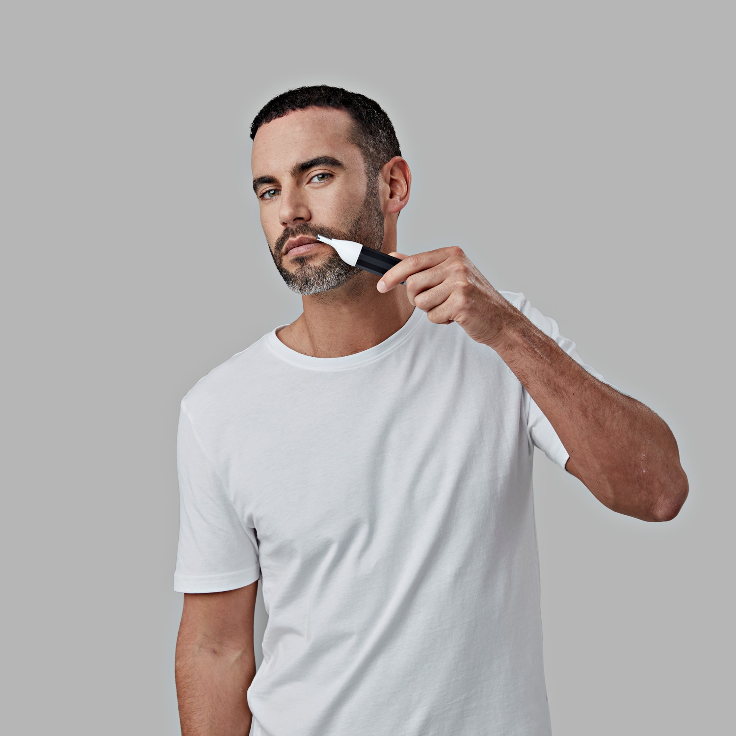 Hombre con camisa blanca recortando la barba con un recortador blanco y negro, mirando al espectador.