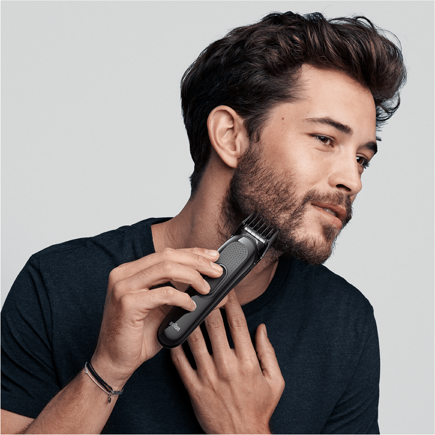 A man shaves his beard with a black Braun trimmer. The man has dark hair and a beard.