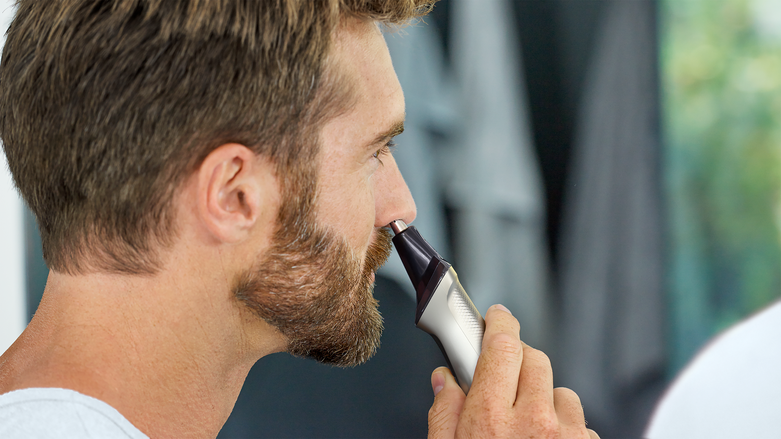 Uomo che si taglia i peli del naso con un trimmer. Ha la barba ed è davanti a uno specchio.
