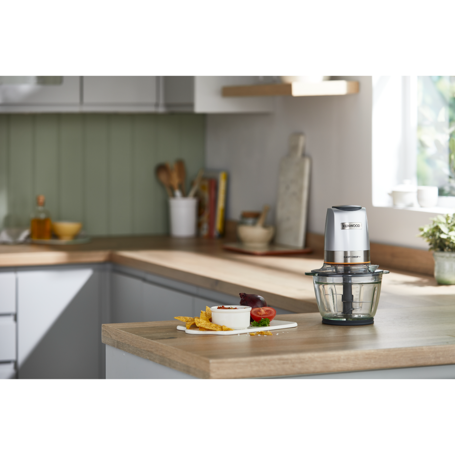 Ahşap bir yüzey üzerinde bir Kenwood mutfak robotu. Bir kase sos, bir domates ve cips görünür.