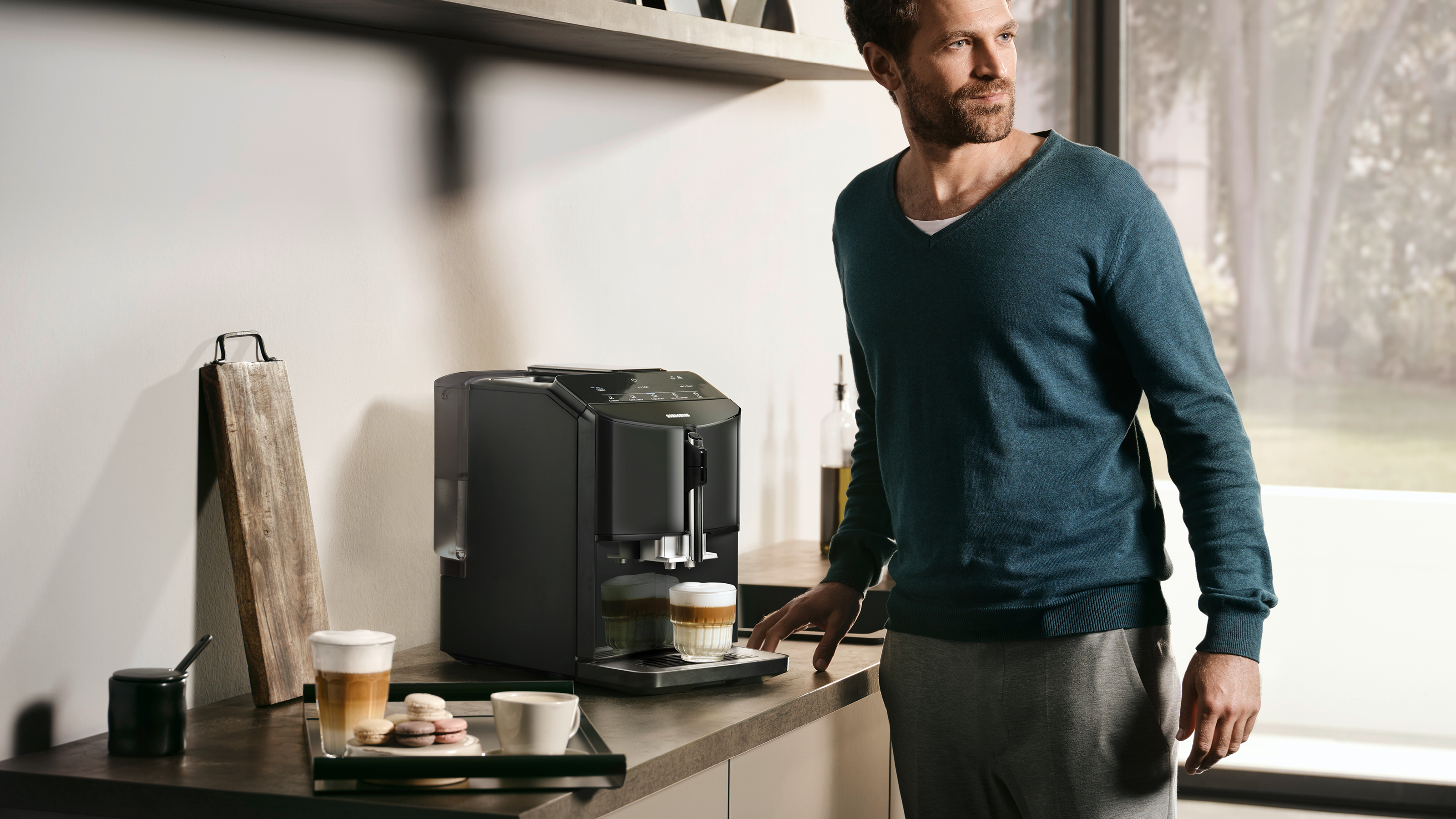 Mężczyzna w turkusowym swetrze stoi przy ekspresie do kawy na blacie z kawą i ciastkami.