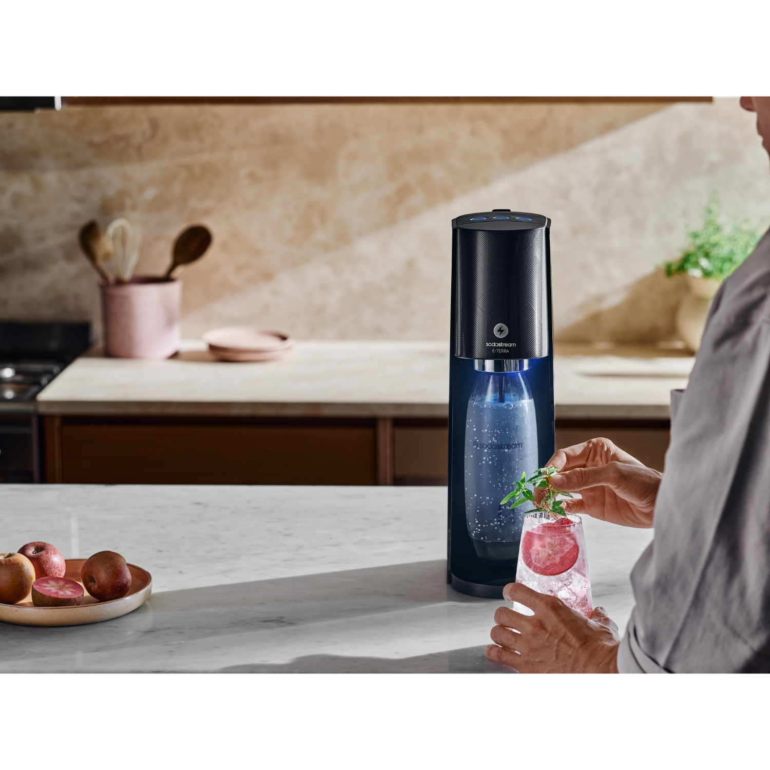 SodaStream-Maschine mit Glas, Person fügt Garnitur auf weißer Arbeitsplatte hinzu.