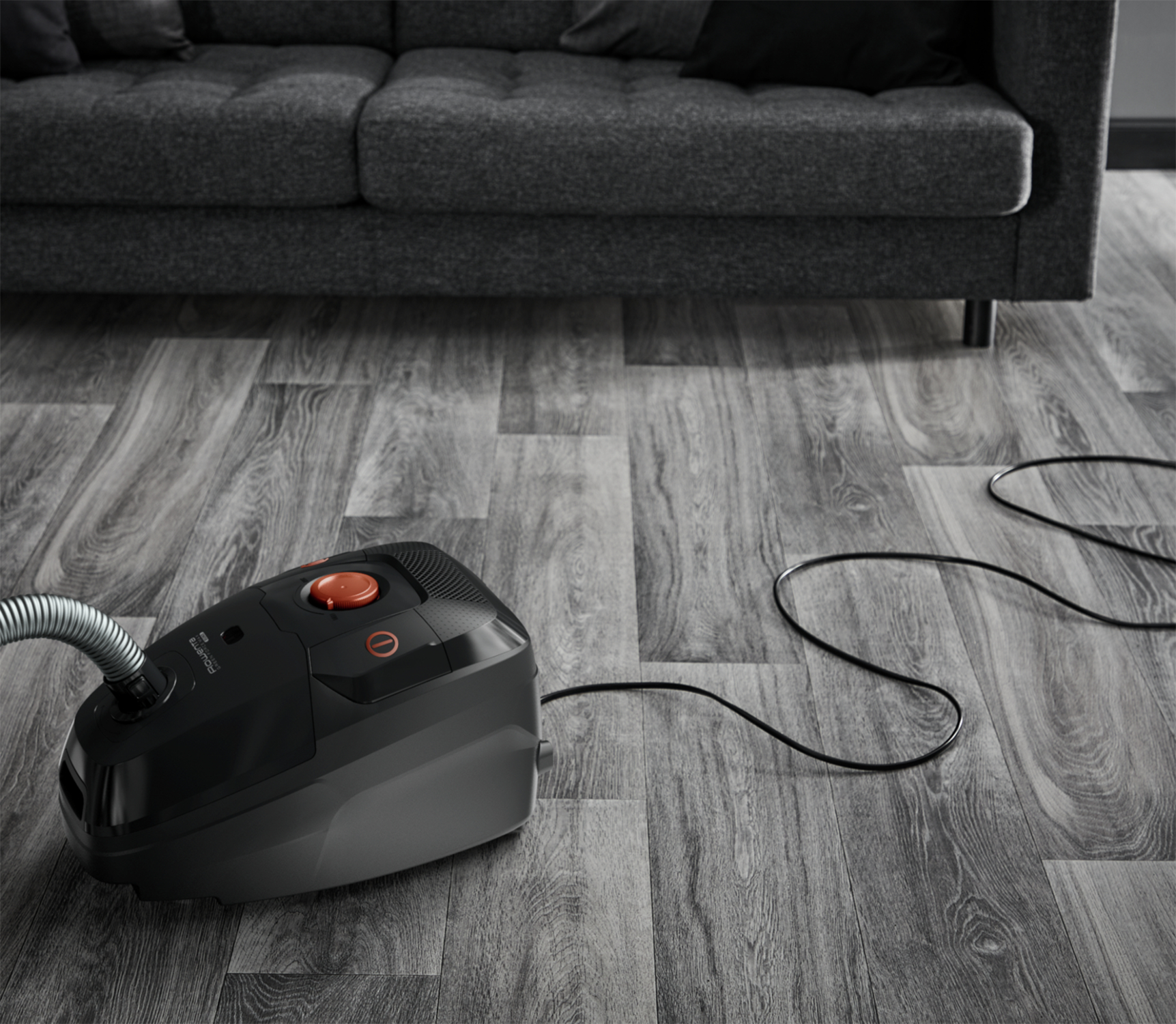Czarny odkurzacz na drewnianej podłodze obok sofy. Widoczny kabel zasilający.