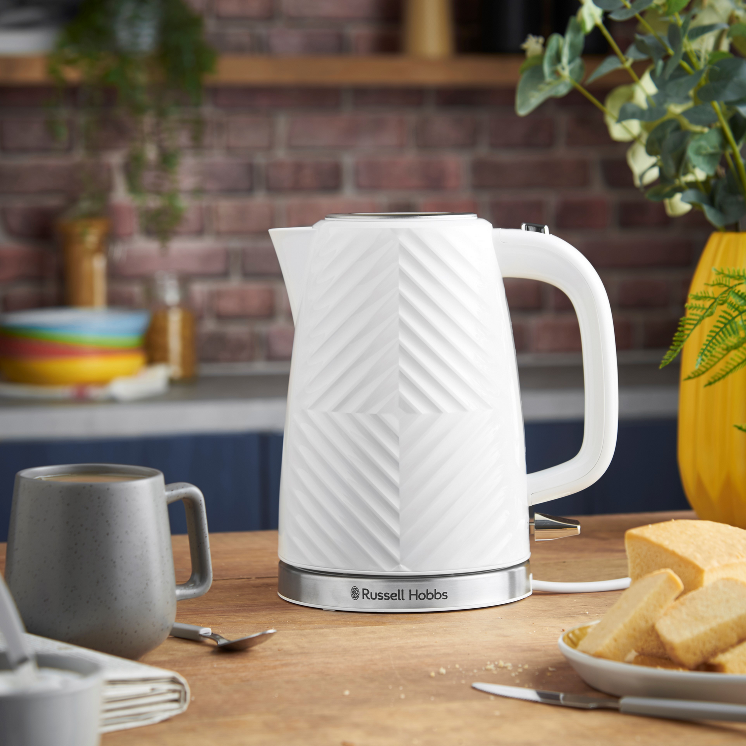 Weißer Russell Hobbs Wasserkocher mit geometrischem Design, neben einer grauen Tasse und Brot.