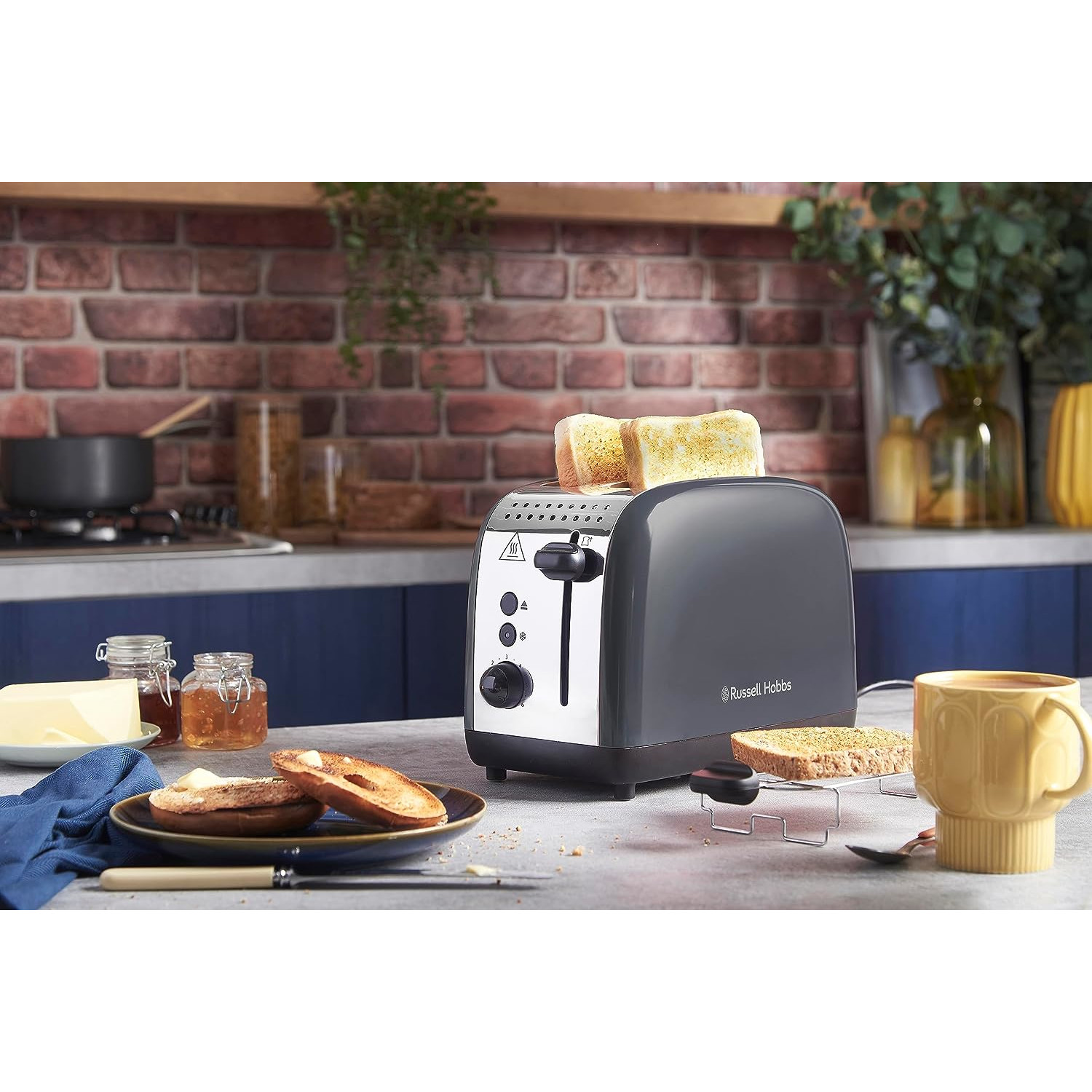 Ein Toaster mit Toast, Butter und Marmelade. Rechts steht ein gelber Becher. Im Hintergrund eine Backsteinmauer.